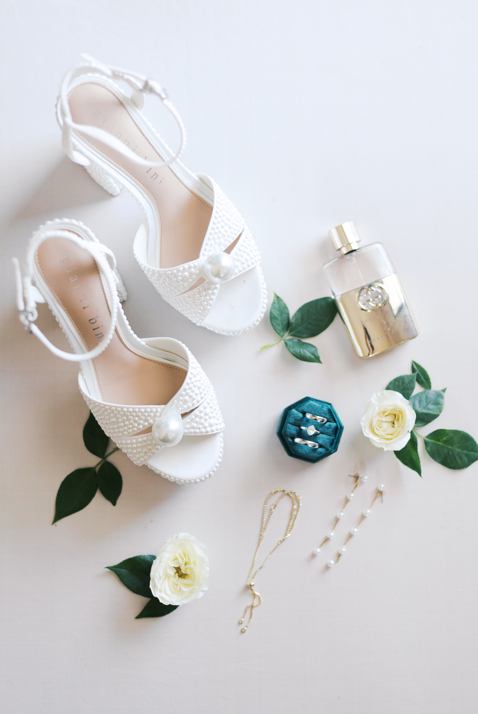 Flat lay of white pearl-studded bridal heels, perfume, gold jewelry, and white roses with greenery on a neutral background.