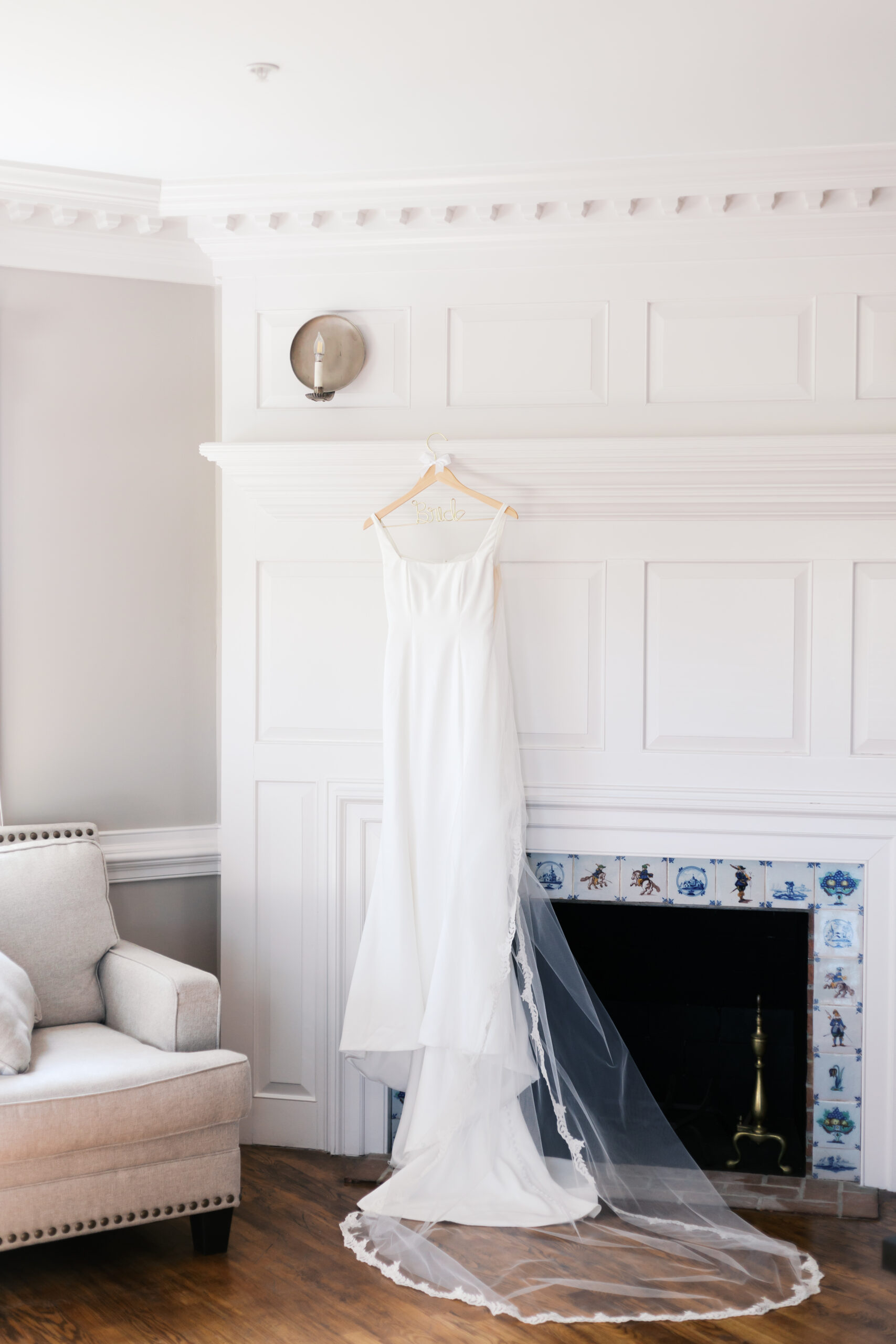 Simple white wedding dress with a long veil hanging on a fireplace mantel with classic white paneling and blue tile accents at Mildale Farm