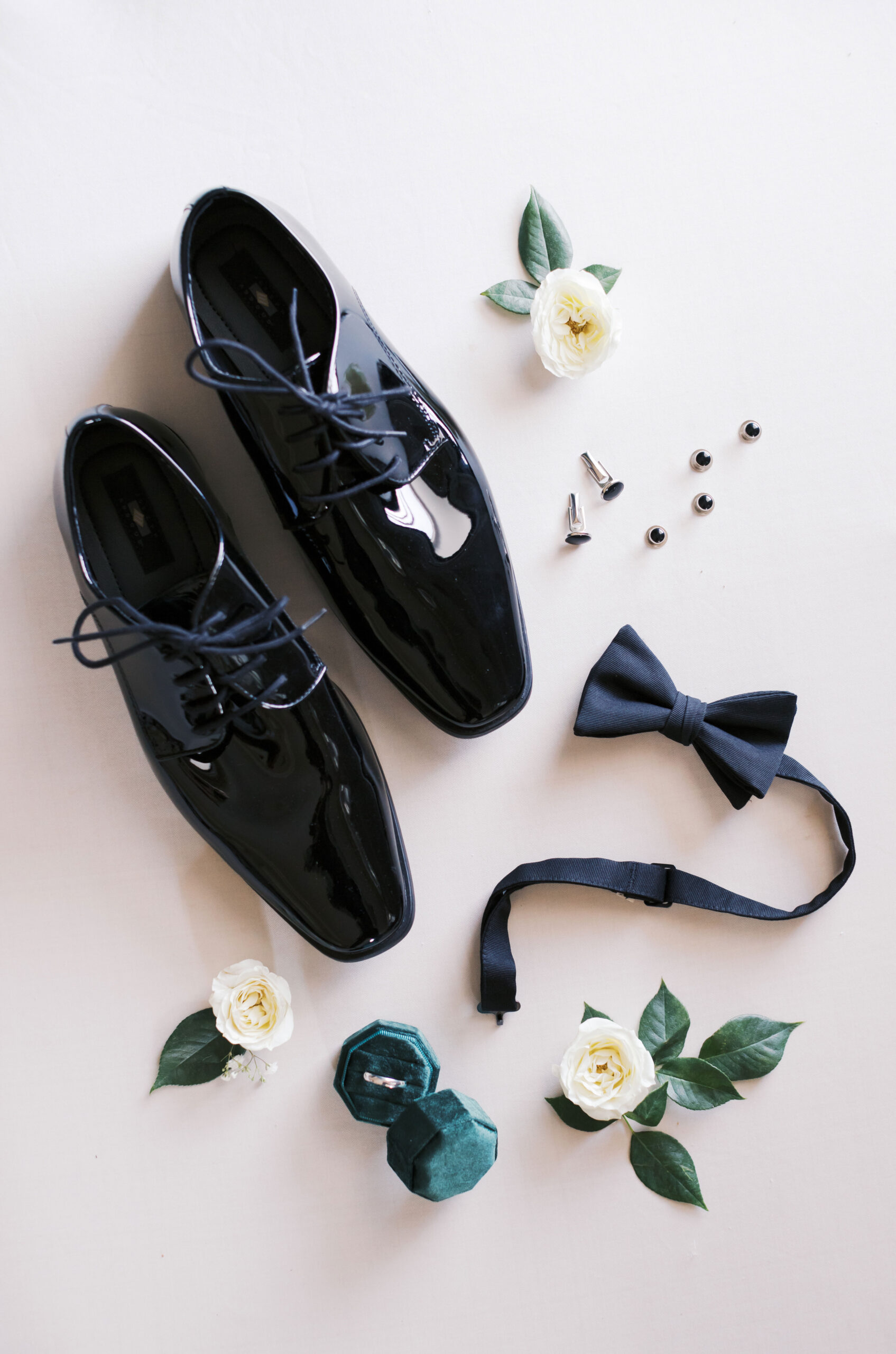 Flat lay of black patent leather dress shoes, black bow tie, cufflinks, wedding band, and white roses with greenery.