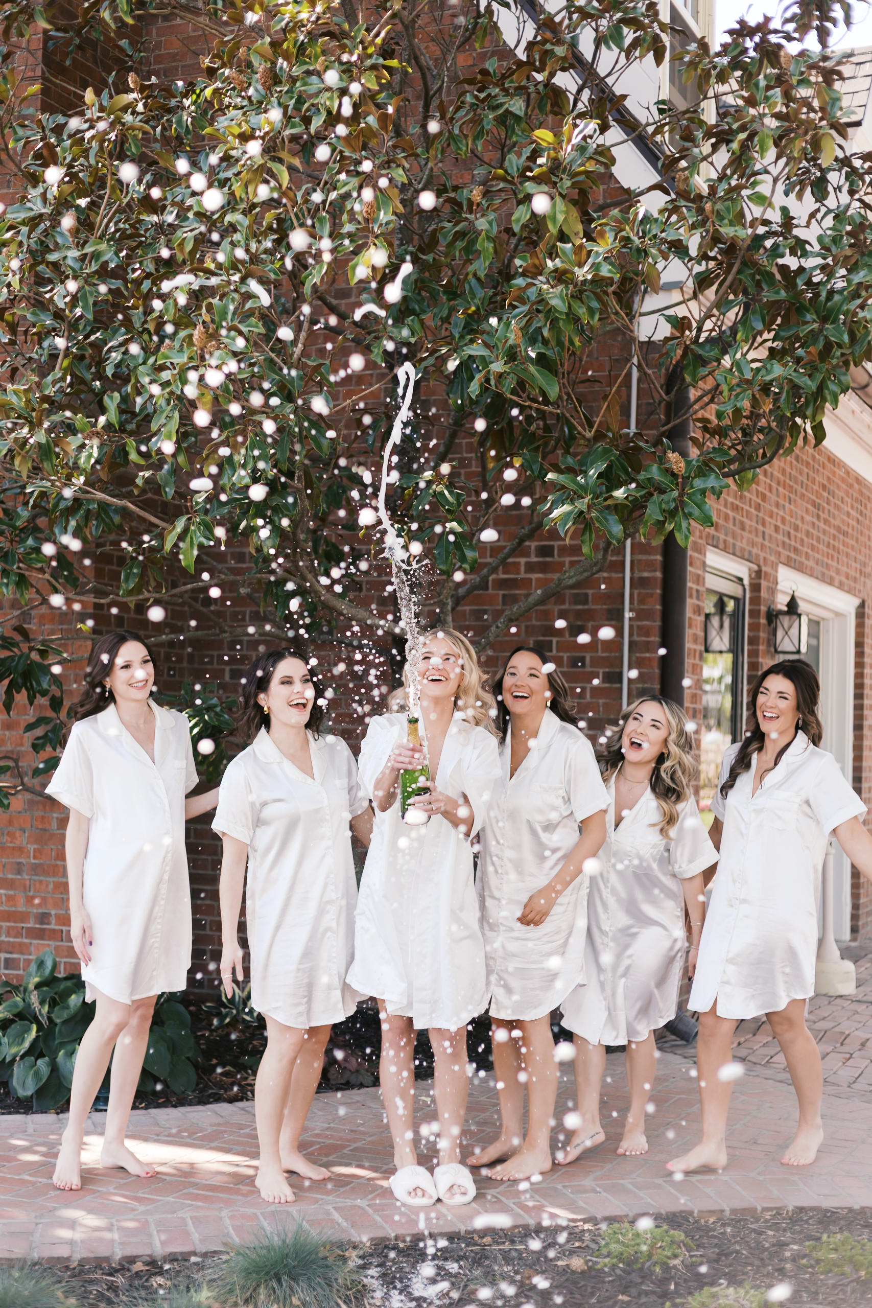 Bride and bridesmaids in white robes popping a bottle of champagne outdoors beneath a tree with champagne spray mid-air.