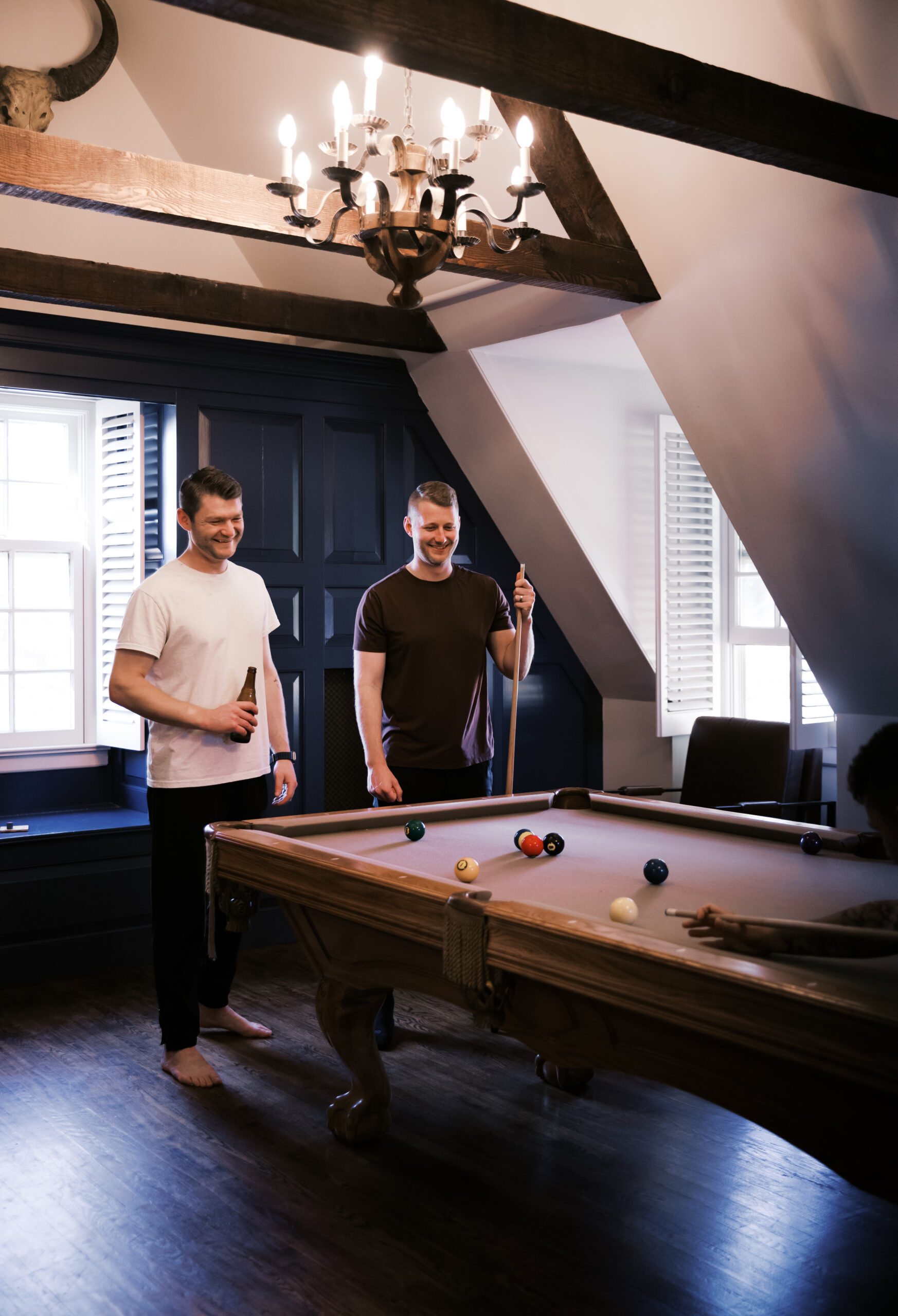 Two groomsmen in casual shirts playing pool in a dark wood and navy-paneled game room with a rustic chandelier at Mildale Farm