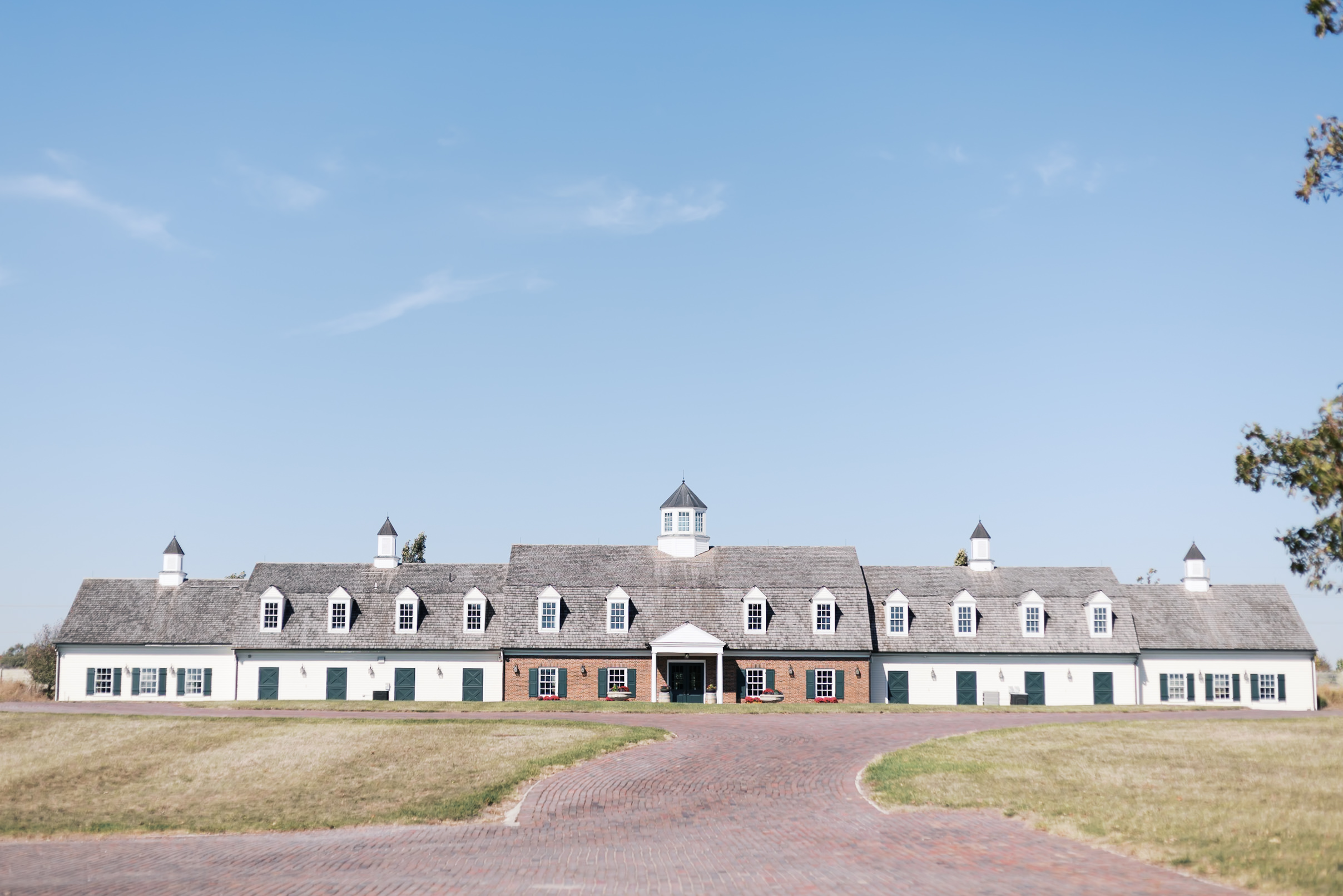 Wide view of Mildale Farm’s charming estate building with dormer windows, cupolas, and a brick driveway under a clear blue sky.