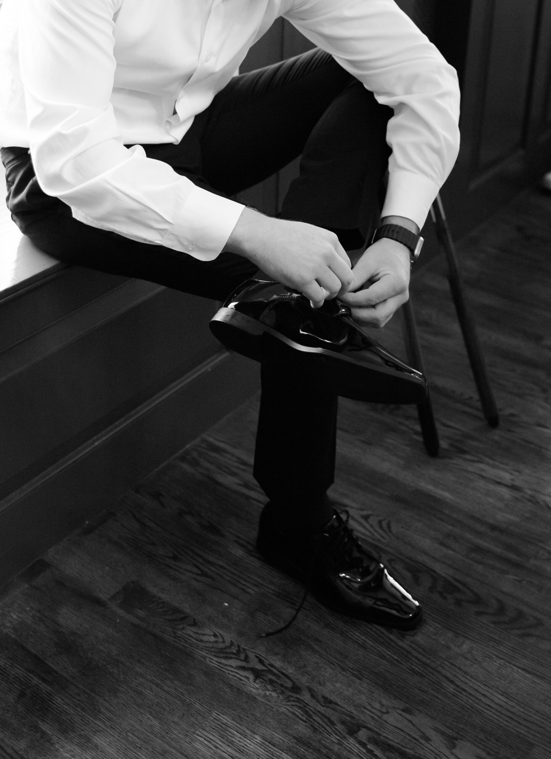 Black and white photo of the groom tying a black dress shoe while seated, wearing a white shirt and dark pants.