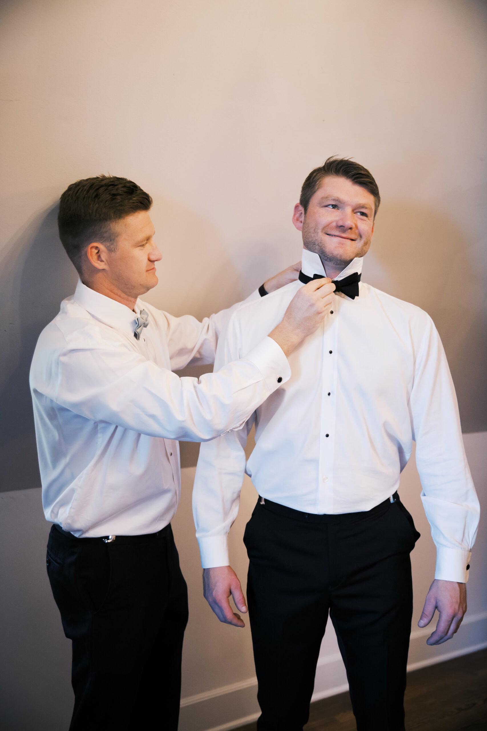 Groomsman adjusts the groom’s black bow tie as they get ready in formal white shirts and dress pants.