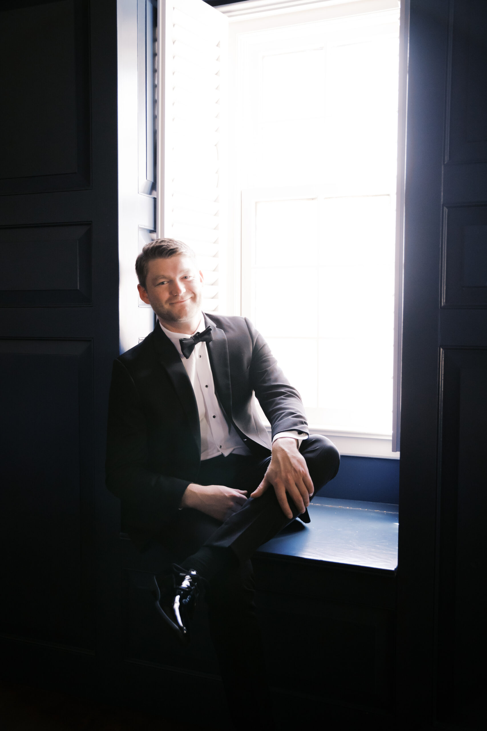 Groom in a black tuxedo sitting by a bright window with navy paneling, smiling while looking relaxed before the ceremony.