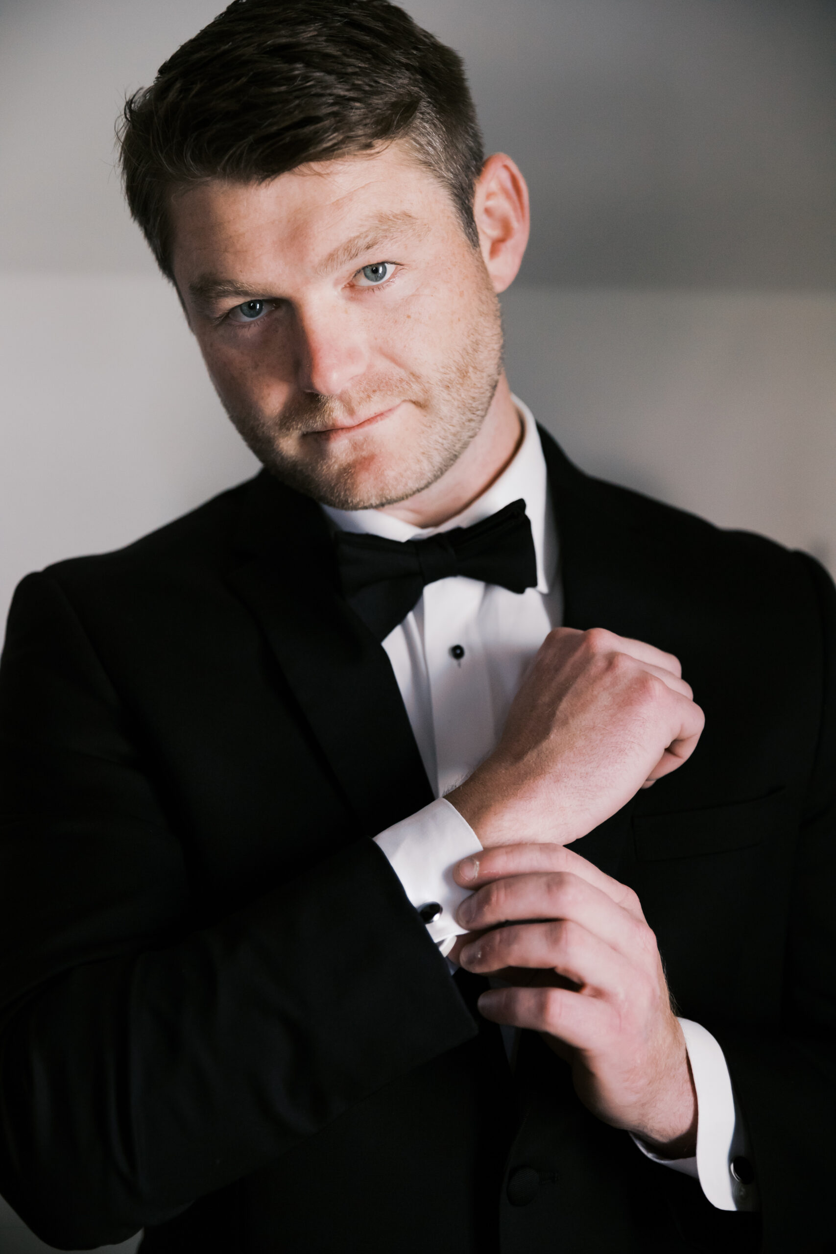 Groom in black tuxedo adjusting cufflink and looking at the camera in a formal pre-ceremony portrait.