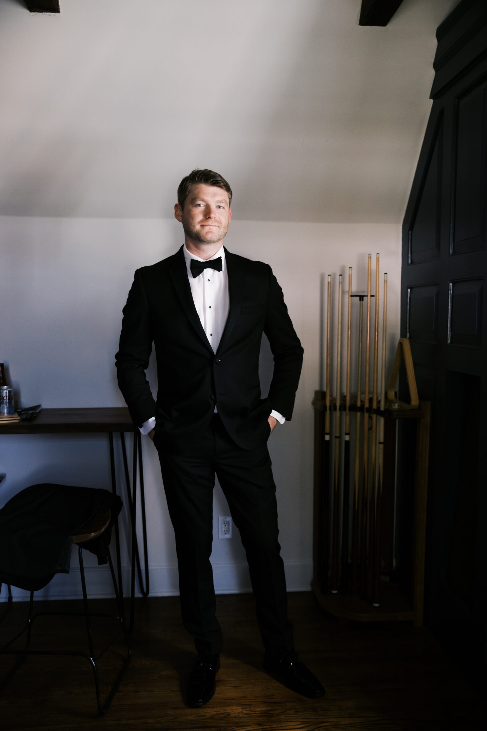 Groom stands confidently in a black tuxedo with hands in pockets inside a stylish room with dark paneling and pool cues.