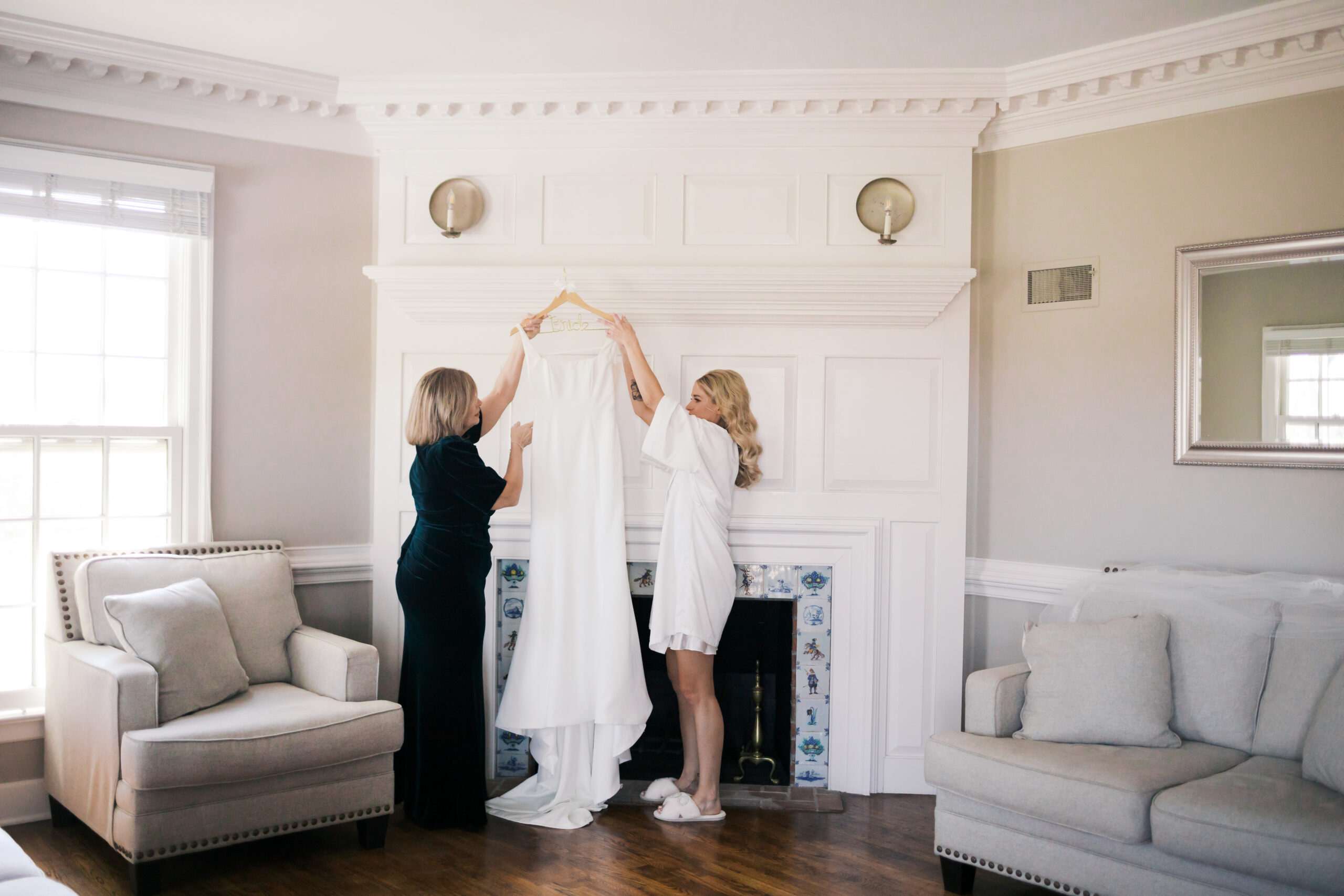 Bride and mother holding up a white wedding dress on a mantel in a soft-lit room with white paneled walls at Mildale Farm.