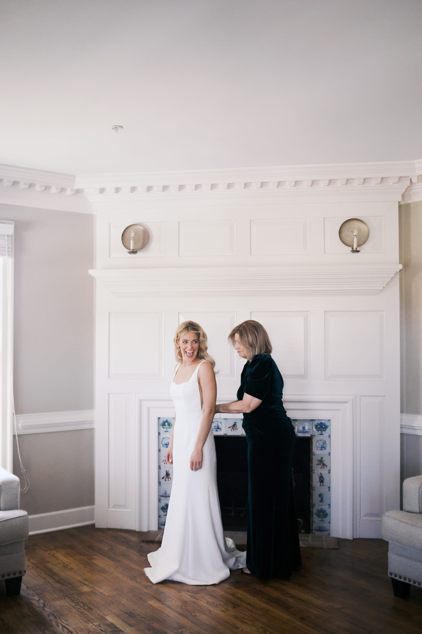 Bride in a simple white gown being helped by her mother during wedding day preparations in a bright indoor space.