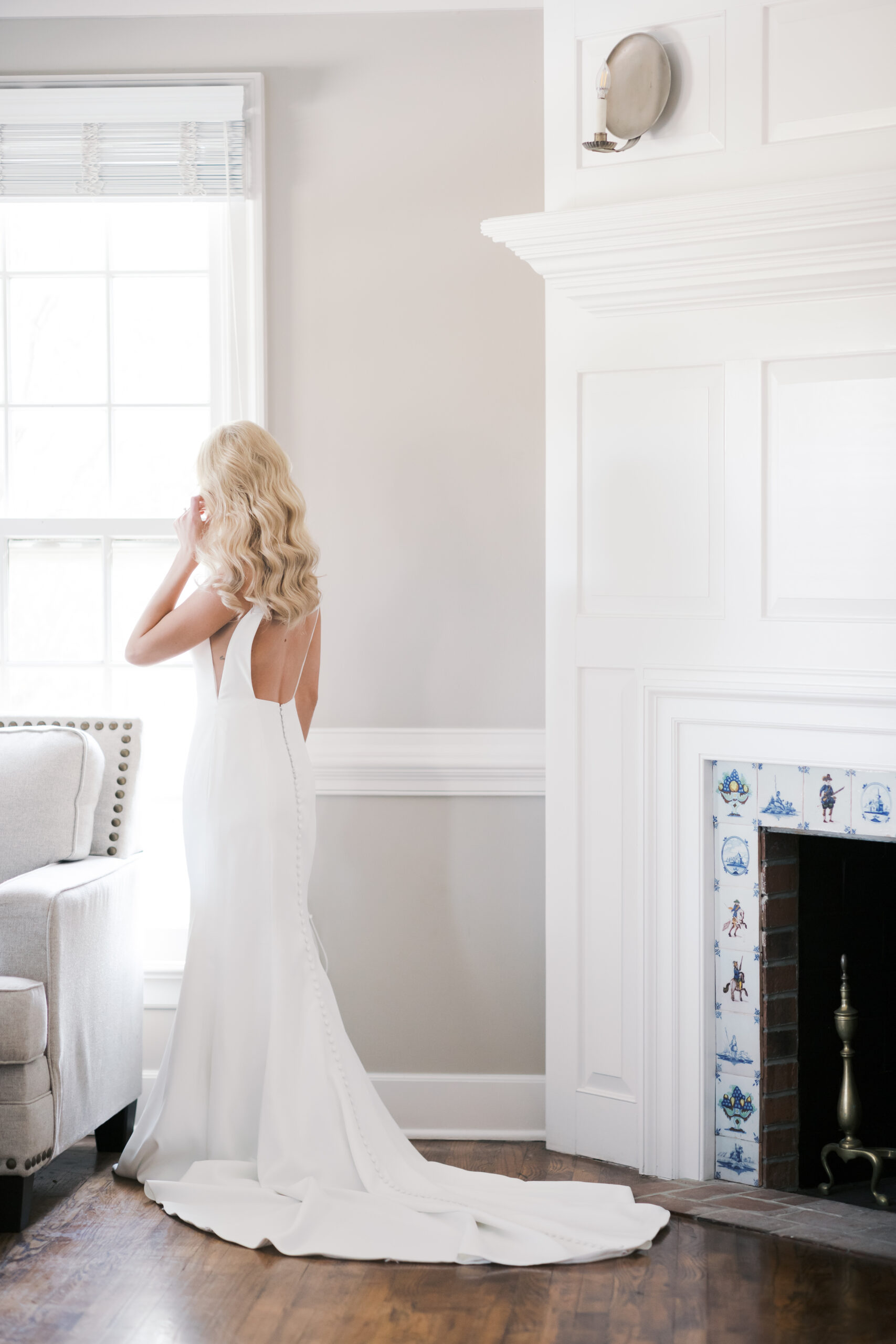 Bride standing by a window in a fitted gown with button detail, facing outside in a soft-lit room with neutral decor.