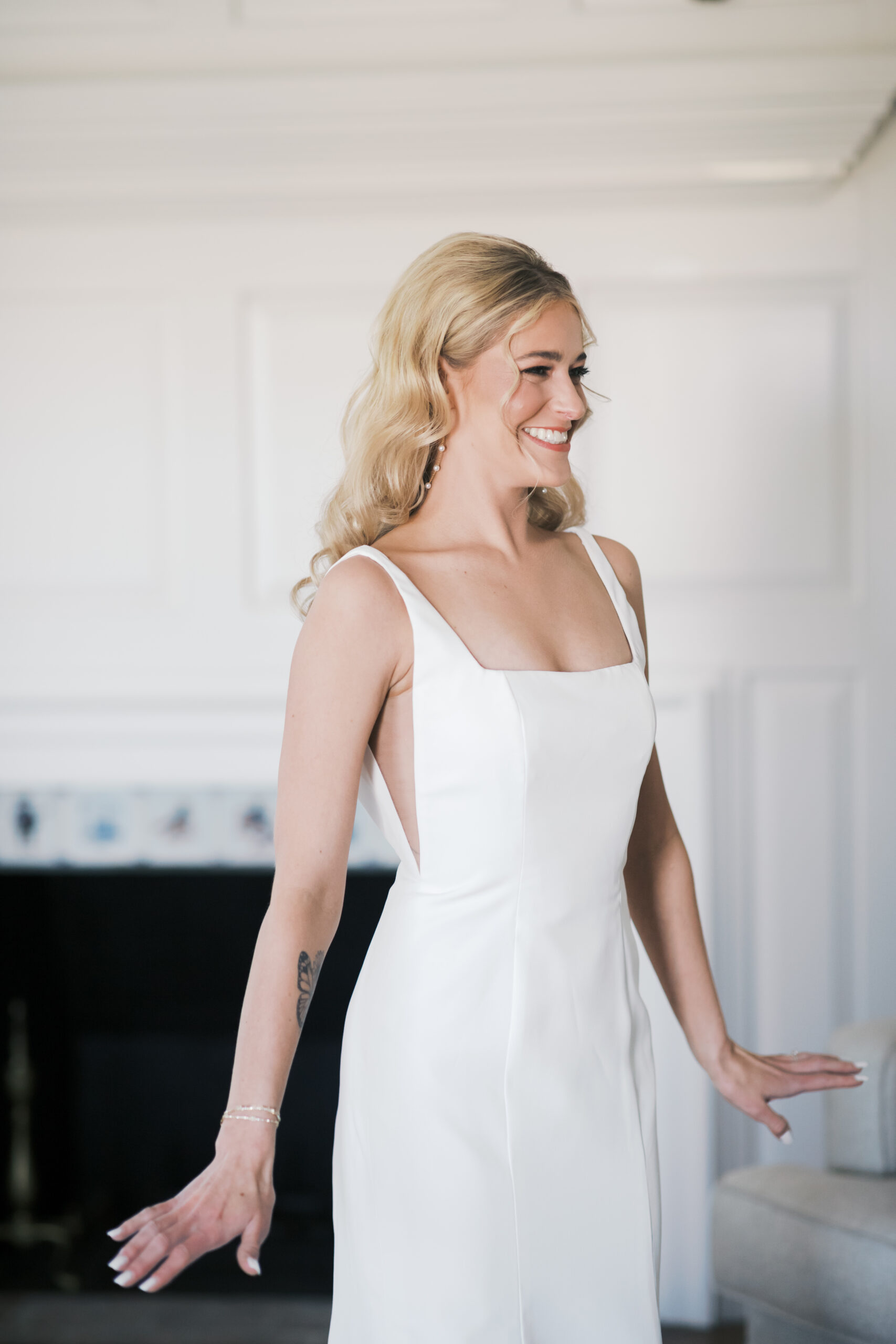 Bride smiling in a simple, elegant white dress with square neckline and long, curled blonde hair before the ceremony.