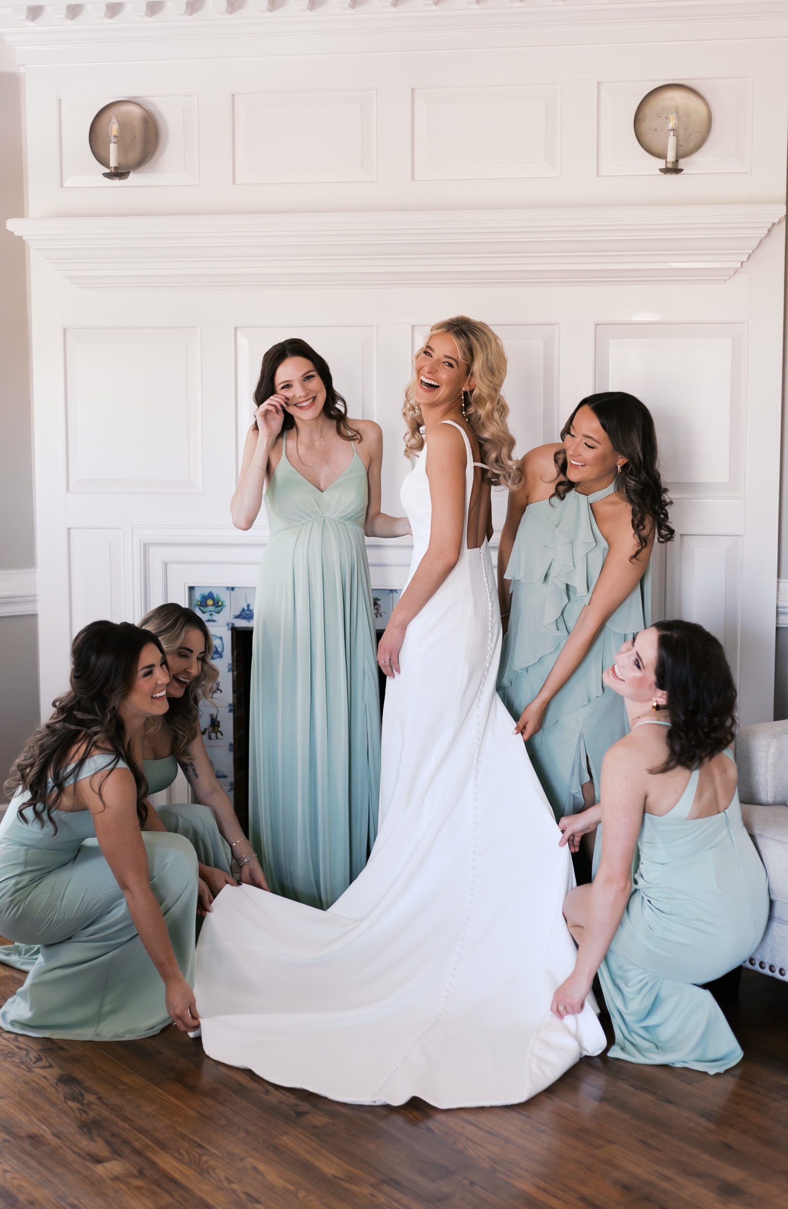 Bride laughing as bridesmaids in sage green dresses gather around to fix her dress train during final getting-ready touches.