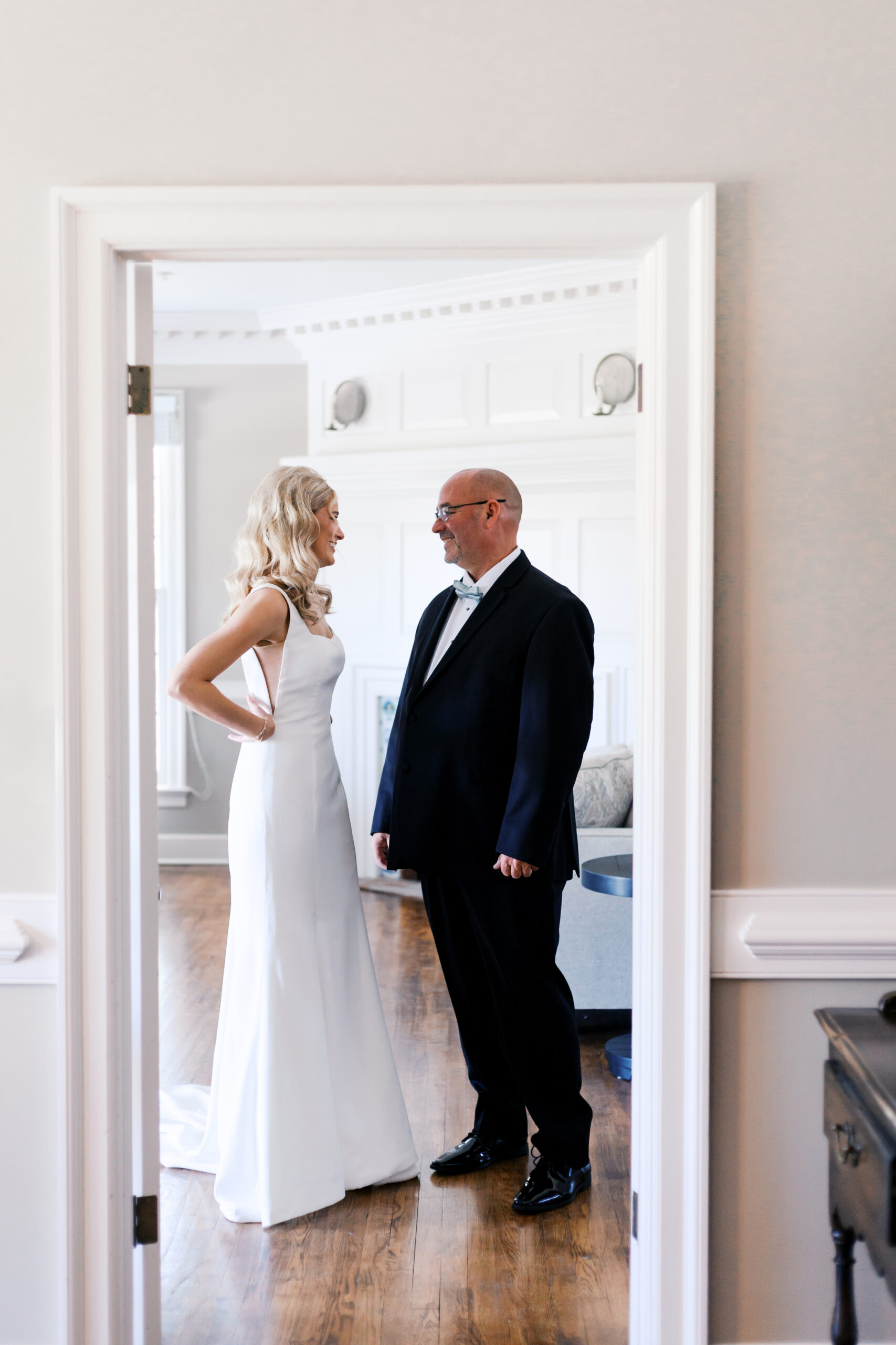 Bride and father smiling at each other across a doorway, framed by elegant white walls and wood floors at Mildale Farm.