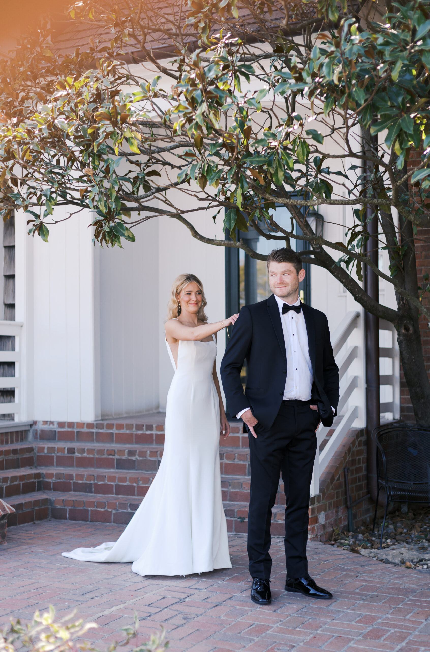 Bride in a fitted white gown taps the groom’s shoulder from behind during their first look beneath a magnolia tree.