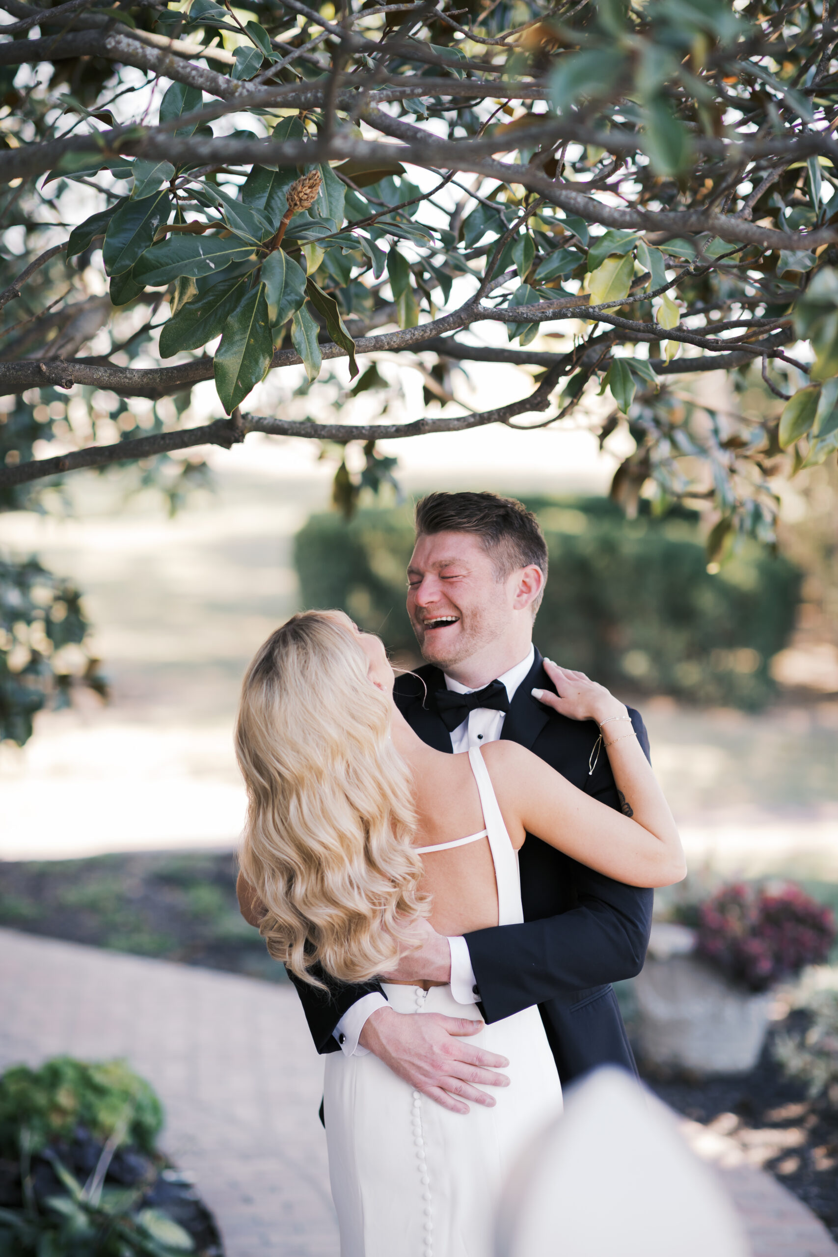 Bride and groom embrace outdoors under leafy branches, smiling and holding each other after their emotional first look.