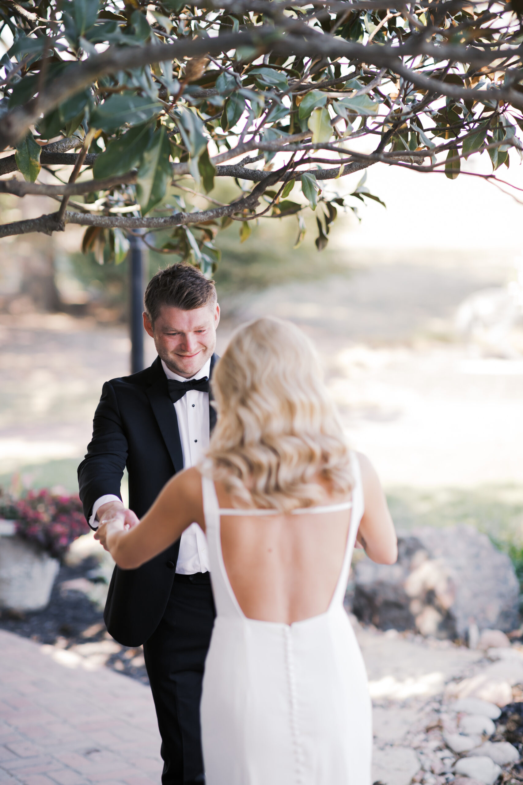 Groom smiles warmly while holding the bride’s hands during their first look outside on a stone path.