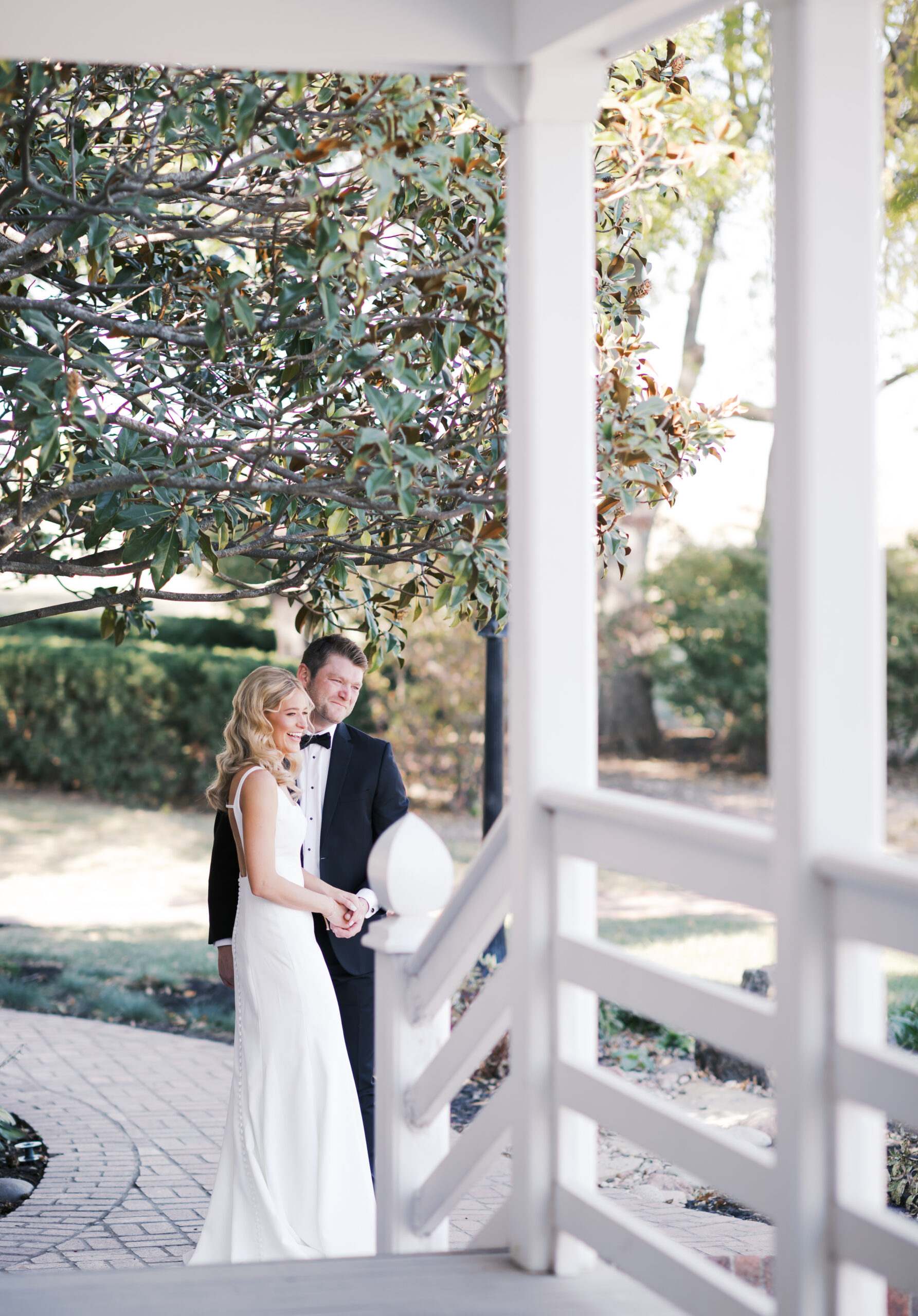 Bride and groom stand side by side on a curved walkway beneath a large magnolia tree, sharing a quiet moment together.