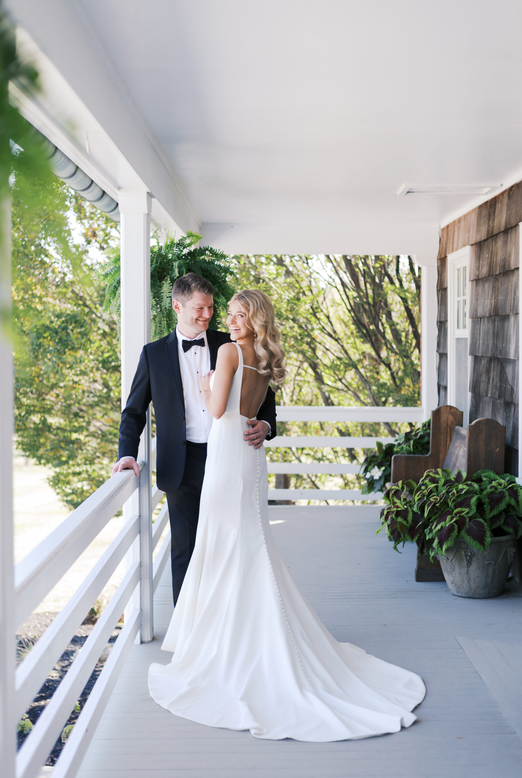 Bride and groom stand close together on a white farmhouse porch, surrounded by greenery and rustic wood siding.