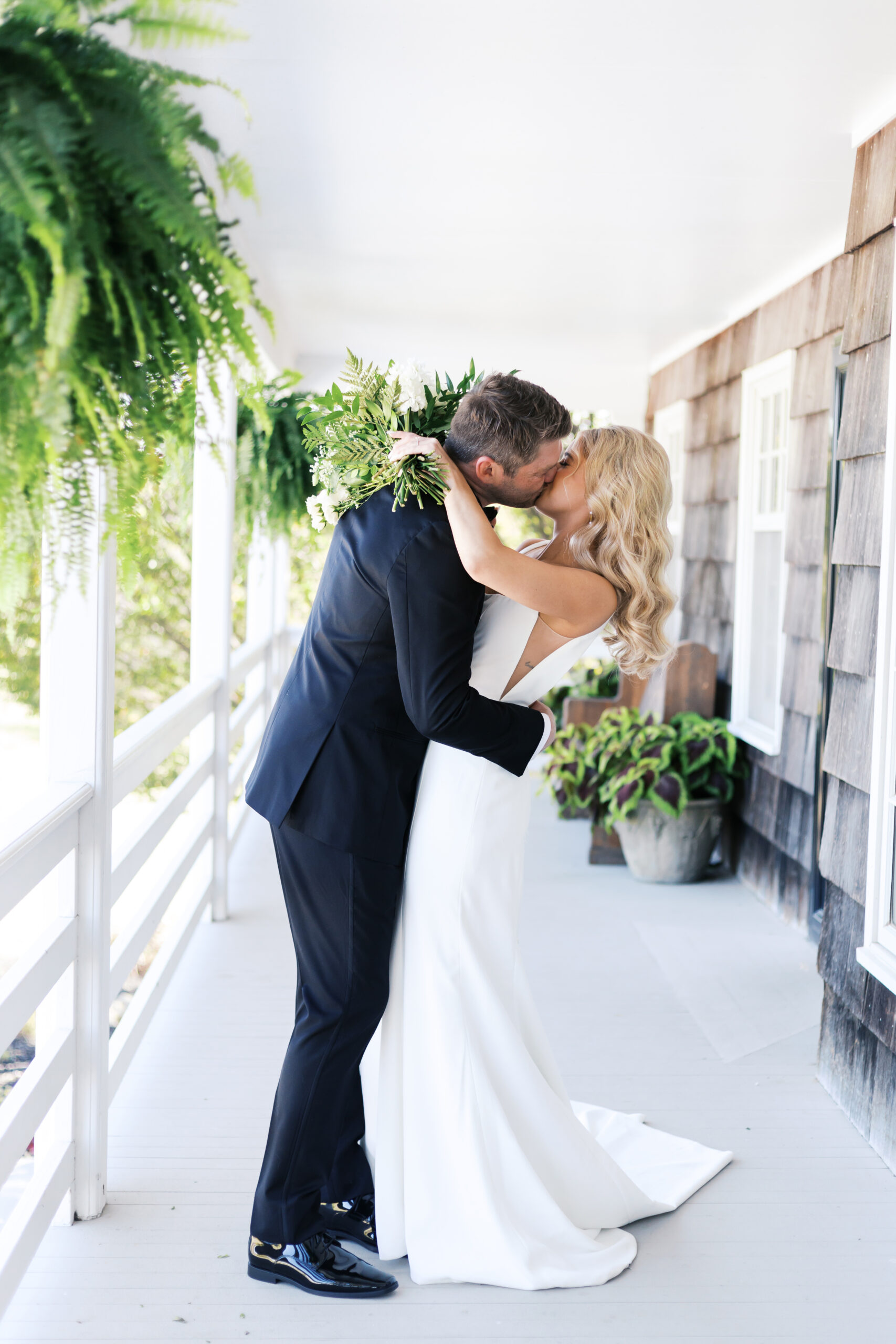 Bride and groom share a kiss on a white farmhouse porch, surrounded by ferns and rustic wood siding, holding a white bouquet.