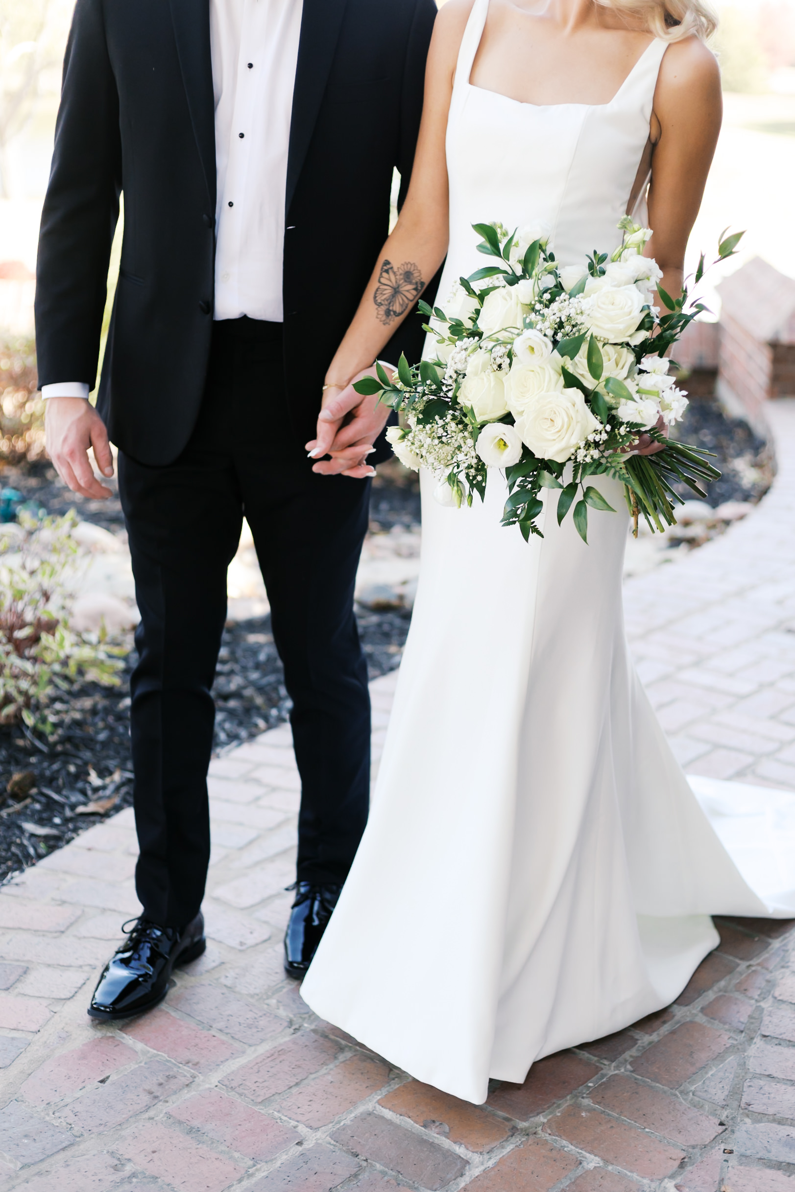 Close-up of bride and groom holding hands, showing the bride’s white bouquet and butterfly tattoo on her arm.