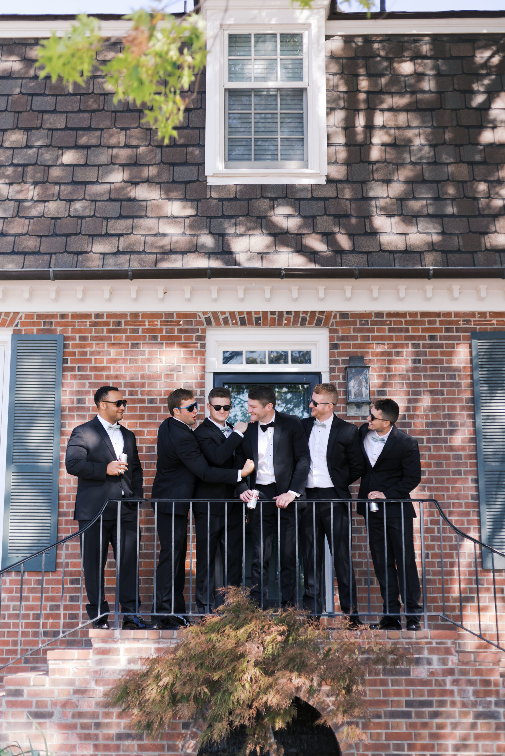Groom and groomsmen in tuxedos stand on a brick balcony with dark shutters, sharing a laugh before the wedding ceremony.