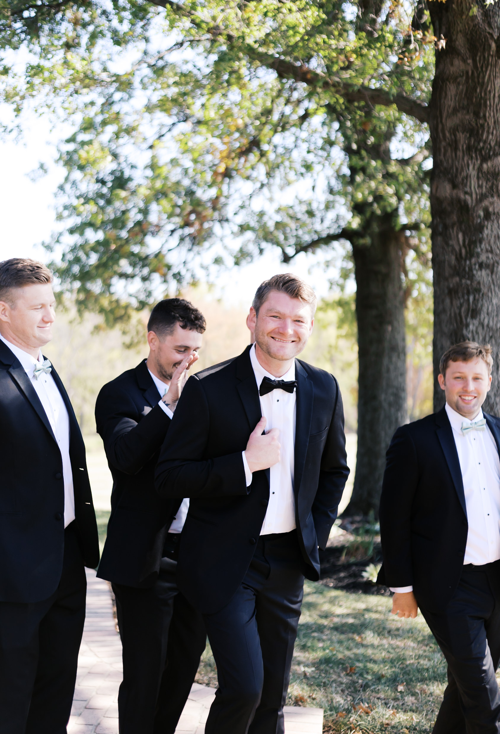 Groom smiles while walking with groomsmen in black tuxedos on a tree-lined path during wedding party portraits.