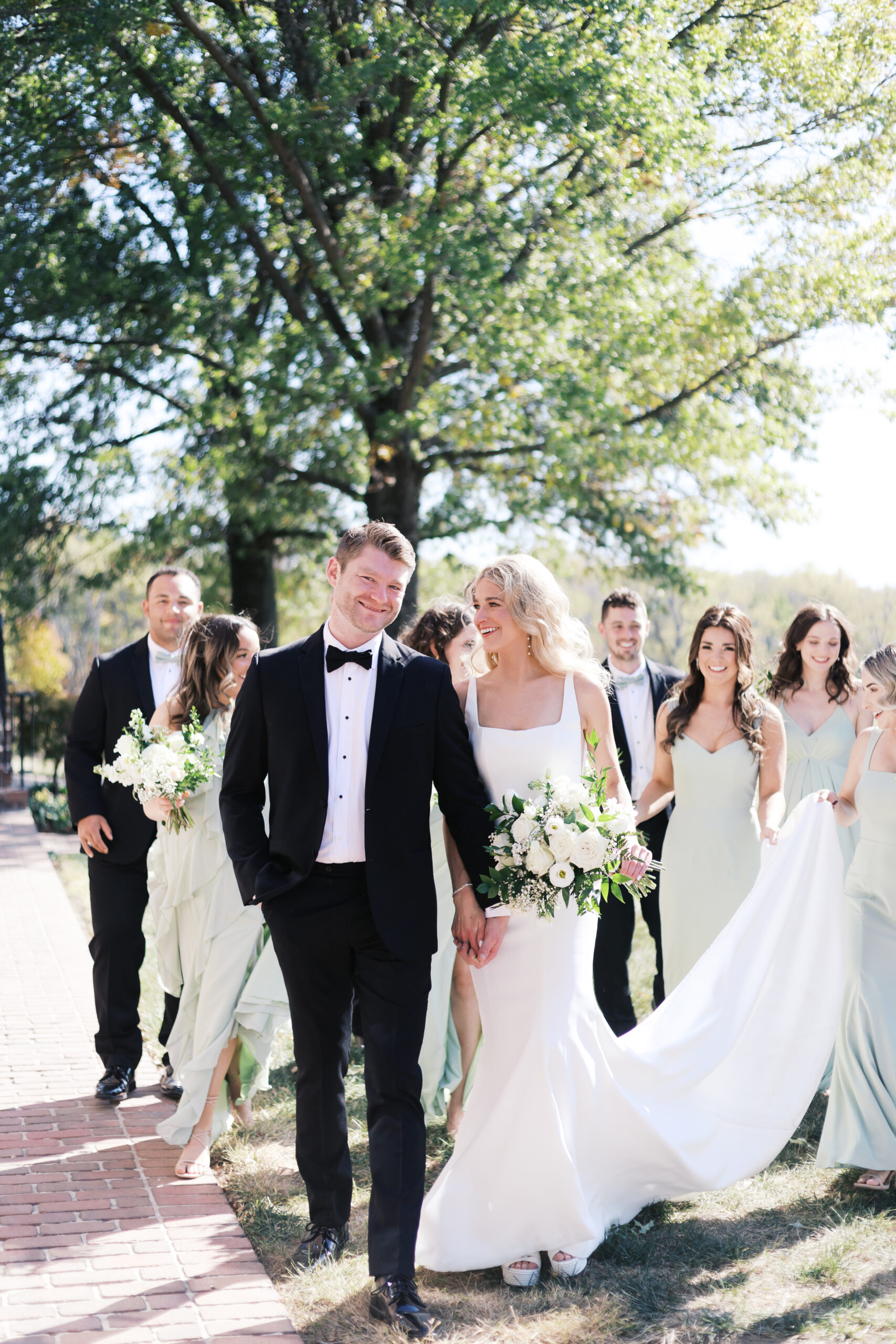 Bride and groom holding hands while walking with bridesmaids and groomsmen on a sunny brick path, all smiling and dressed in formalwear.