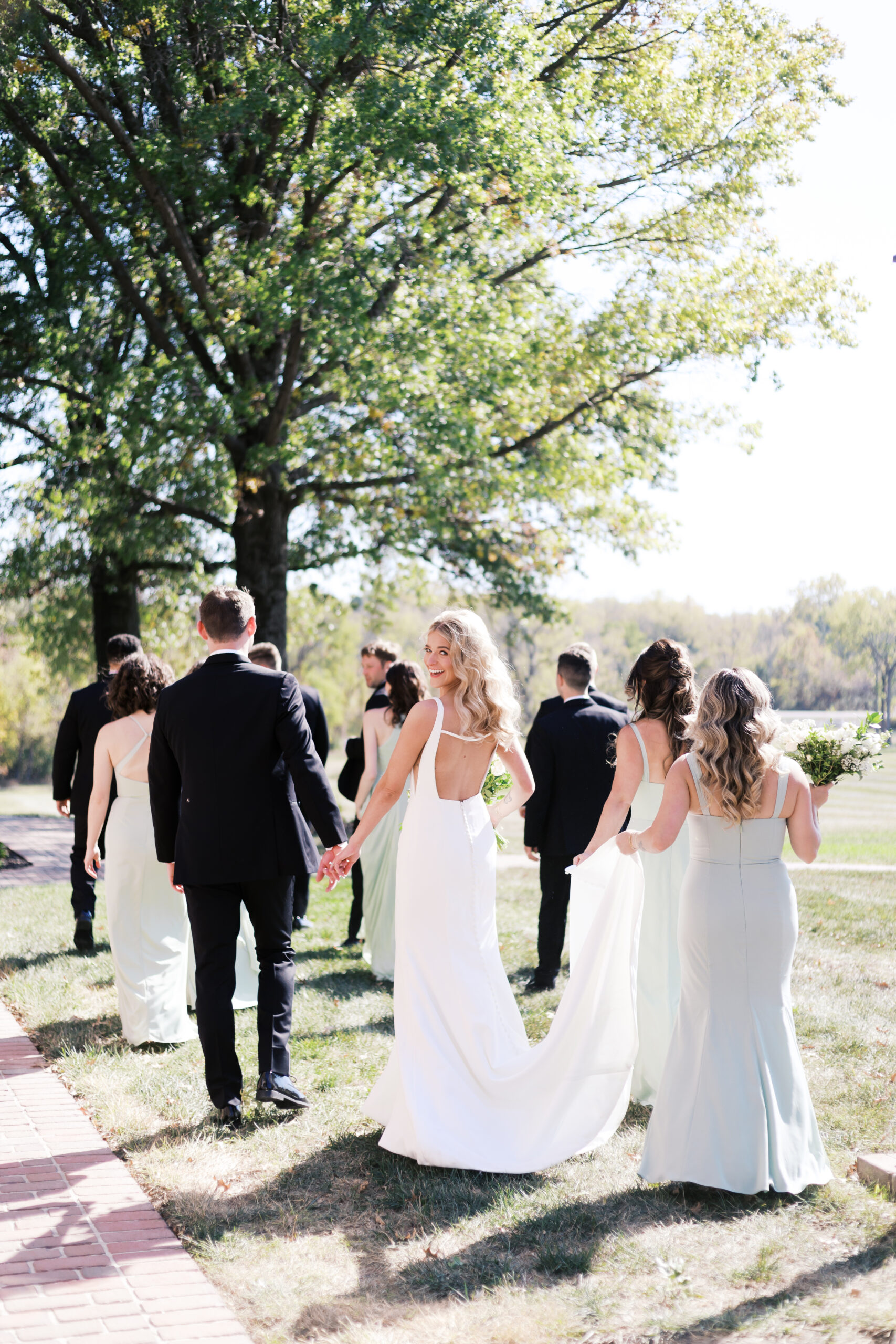 Bride in a backless gown looks over her shoulder while walking hand in hand with the groom and their wedding party across a grassy lawn.