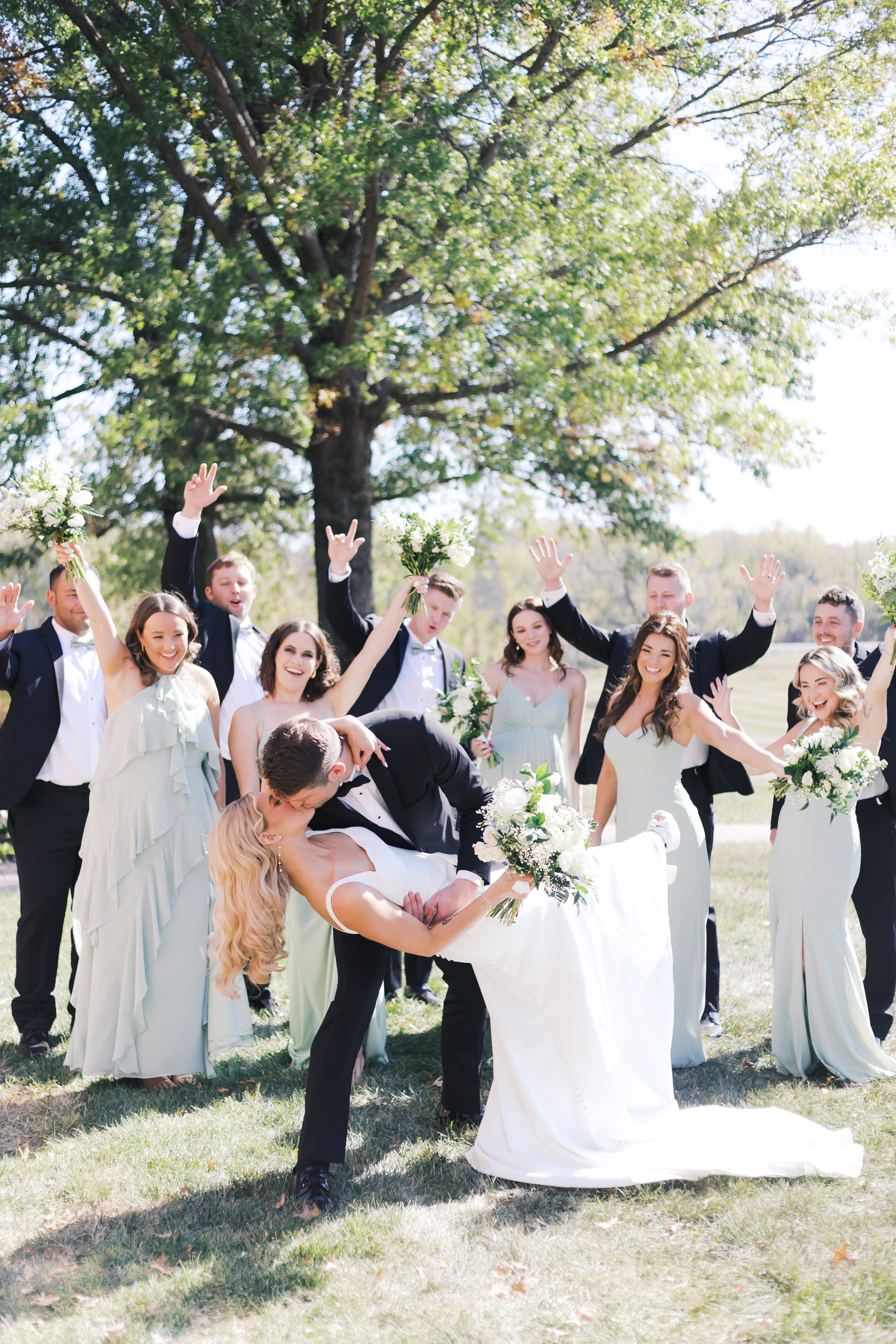 Groom dips the bride for a kiss while wedding party cheers in the background, all dressed in formalwear under a large oak tree.
