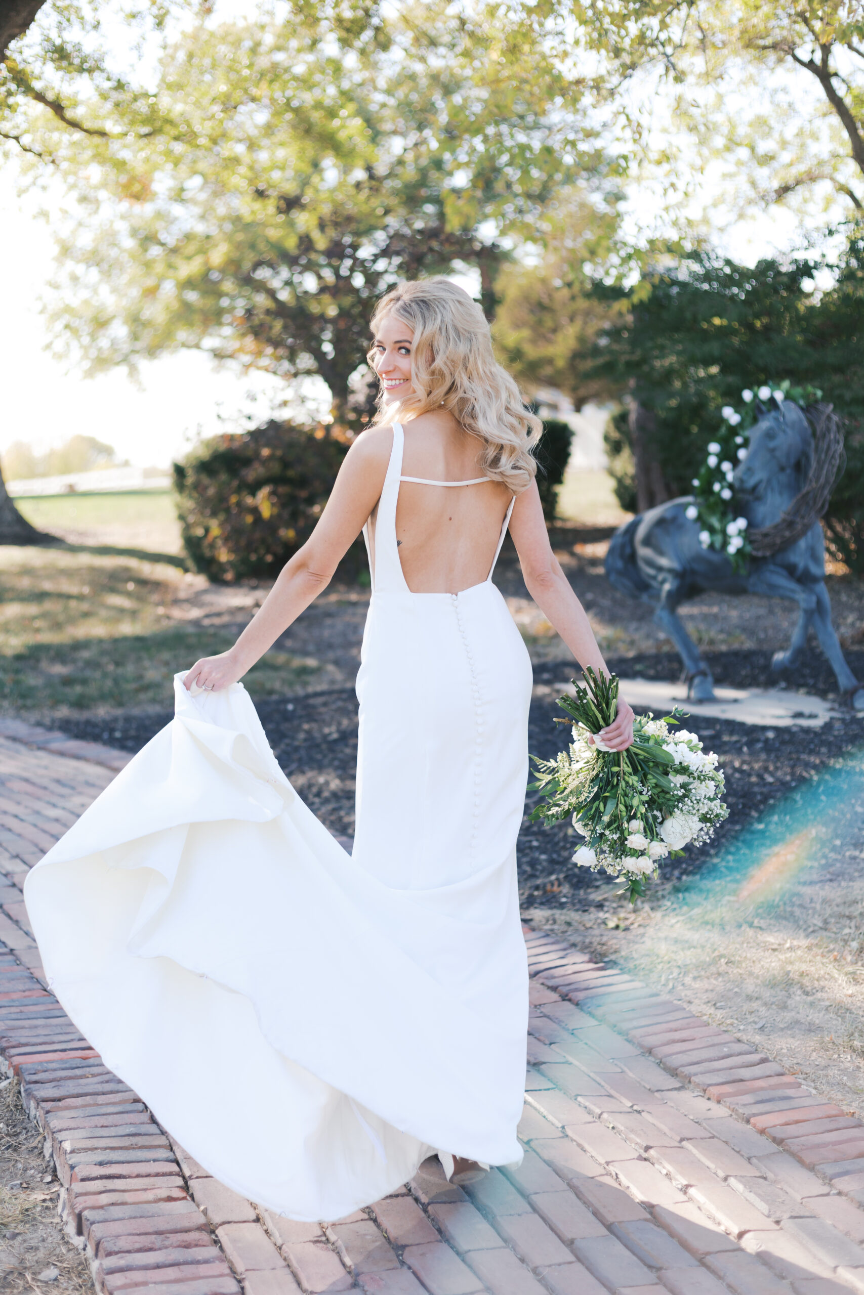 Bride holding a bouquet lifts her train while smiling over her shoulder, walking on a brick path near a horse statue at Mildale Farm.