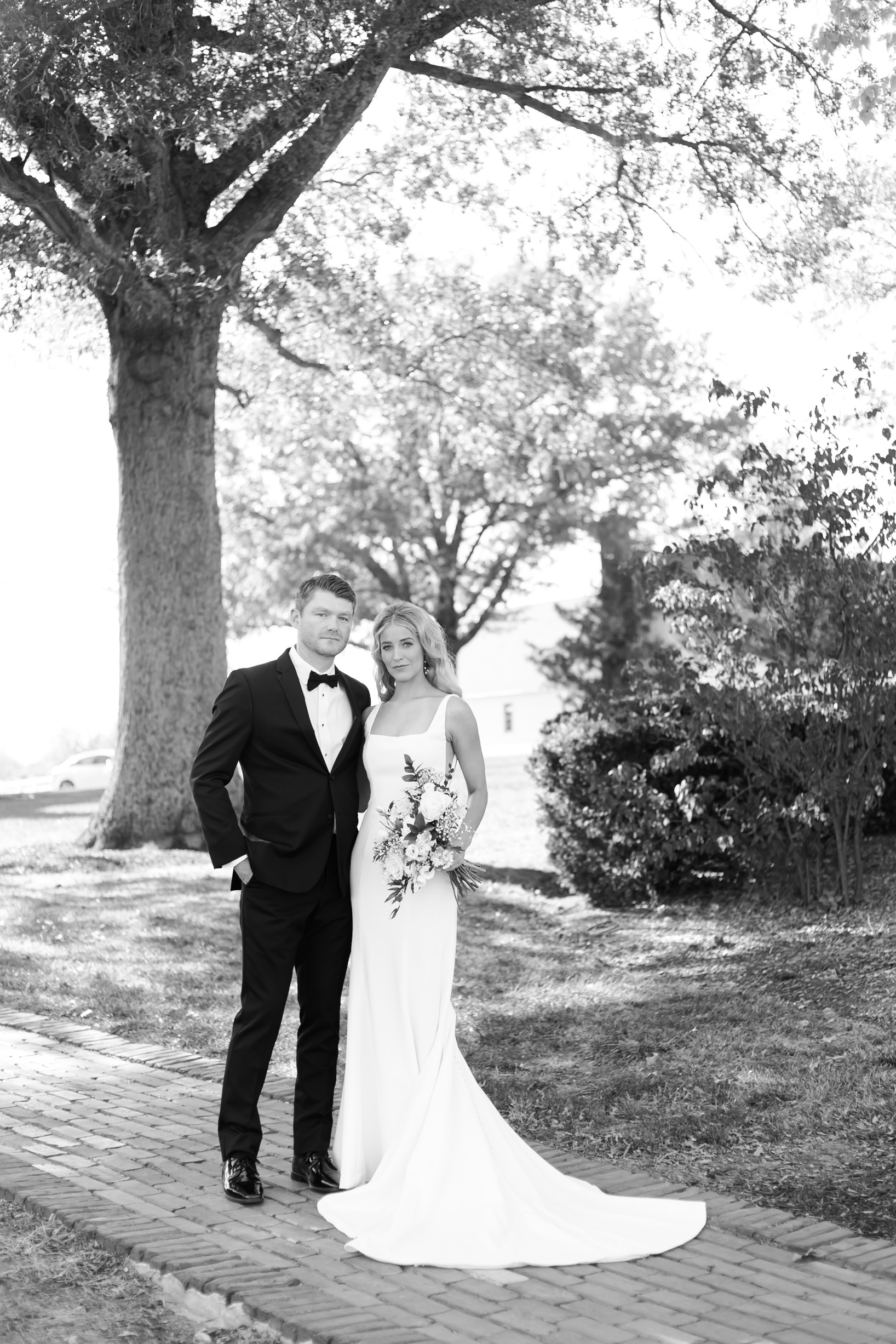 Black and white photo of the bride and groom standing together on a brick path, surrounded by trees and soft afternoon light.