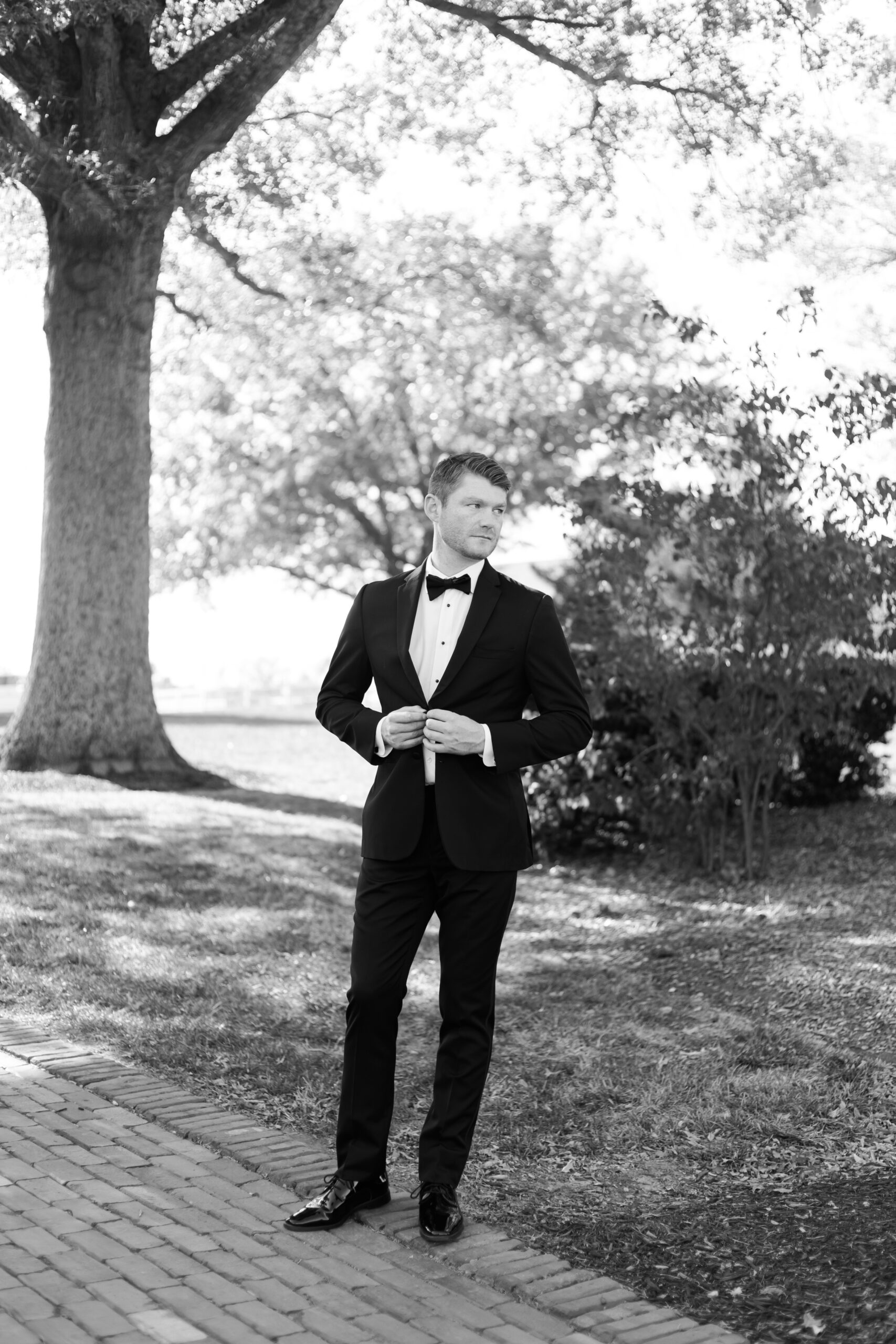 Black and white portrait of groom in a black tuxedo buttoning his jacket under a tree-lined path before the ceremony.