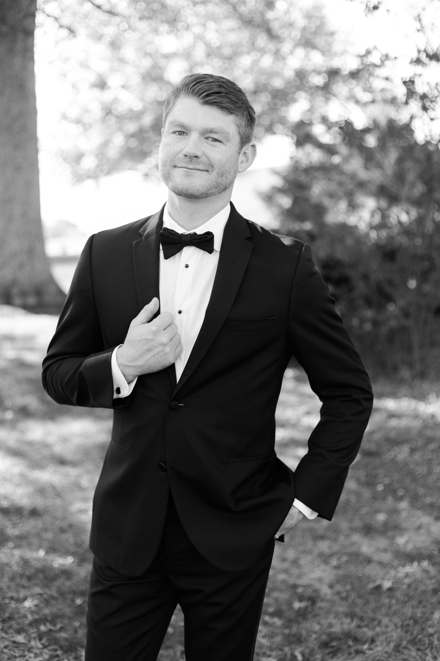 Black and white close-up of the groom smiling slightly, standing in a tuxedo under dappled sunlight.