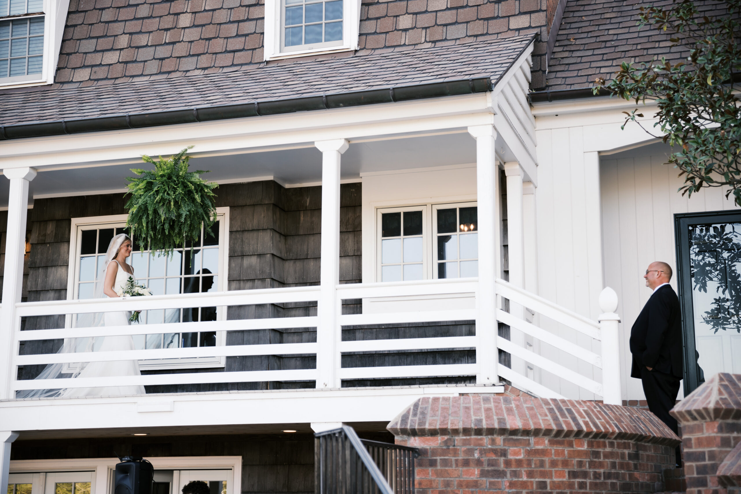 Bride in a white gown walks toward her father on the white upper balcony of a rustic farmhouse.