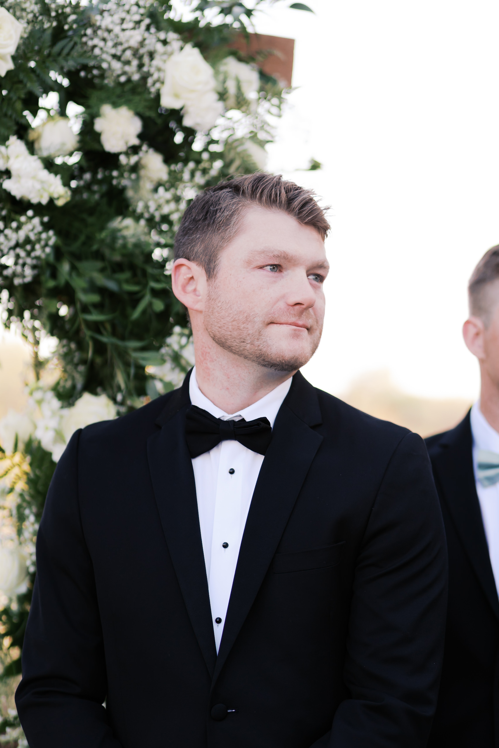 Groom in a black tuxedo standing beneath a white rose floral arch, watching the ceremony begin at Mildale Farm.