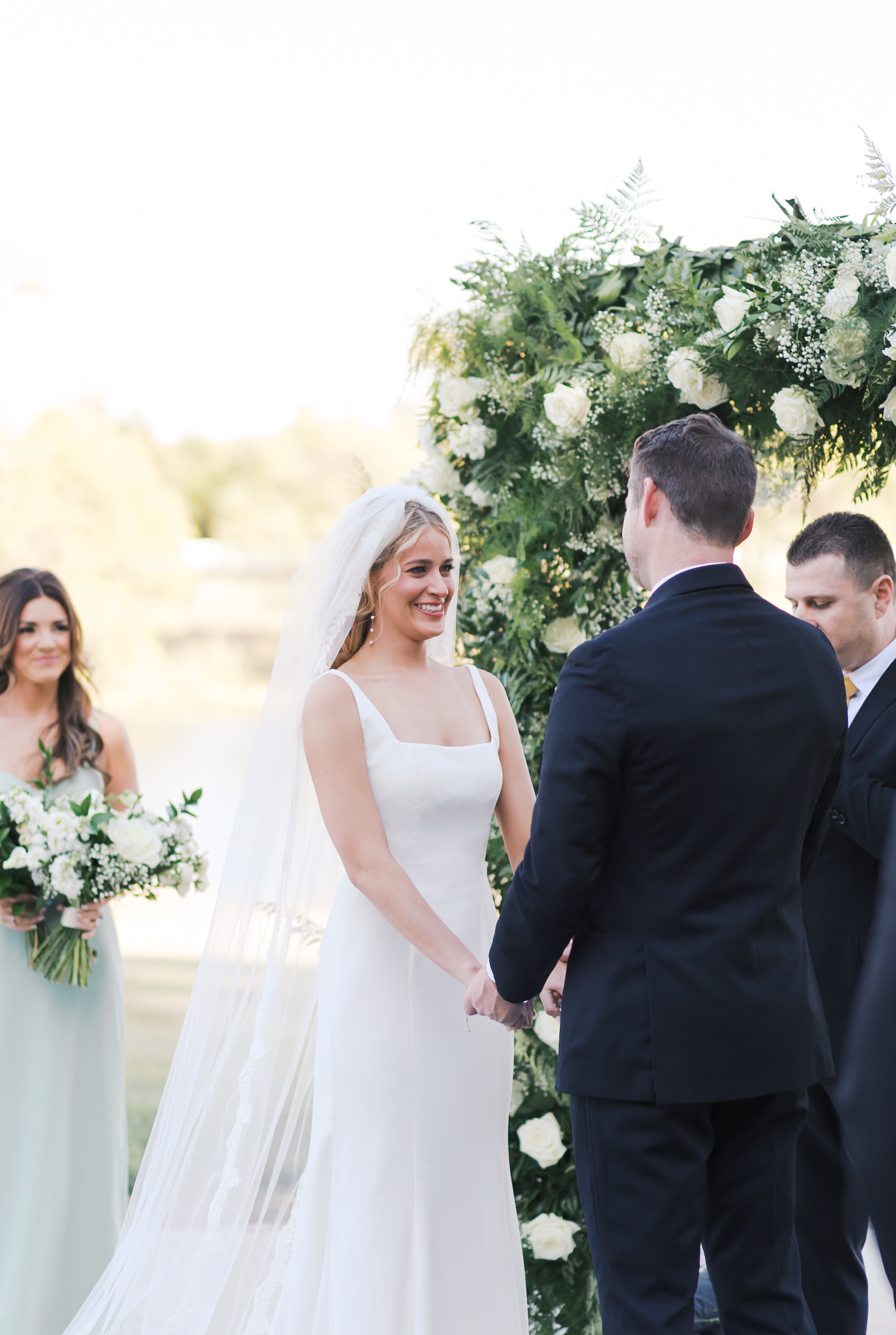 Bride and groom smile as they hold hands beneath a white rose floral arch during their outdoor ceremony at Mildale Farm.
