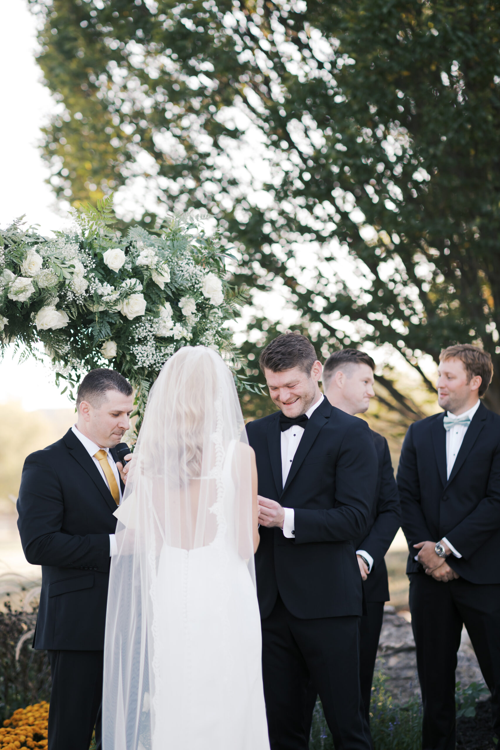 Groom places wedding band on bride’s finger during outdoor ceremony, as the officiant reads beside a white floral arch.