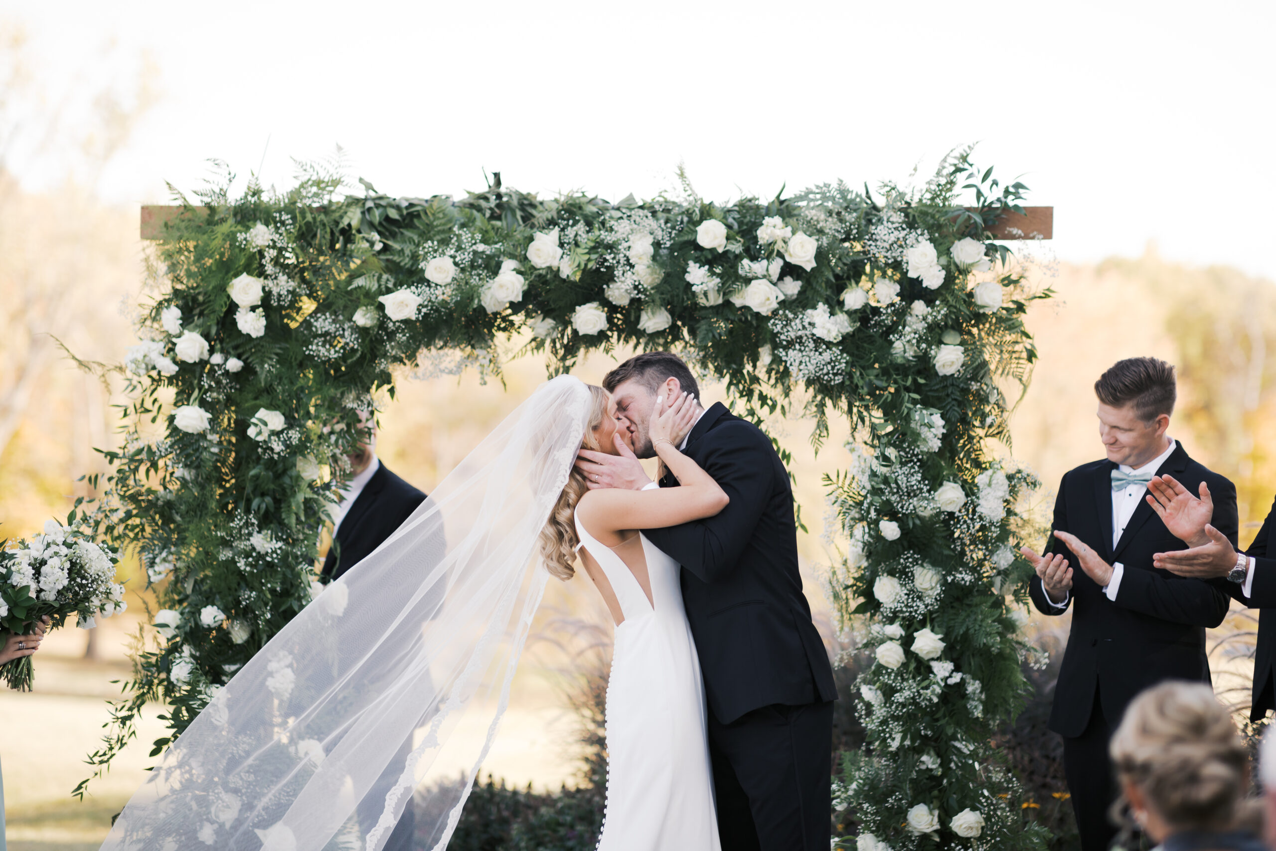 Bride and groom share their first kiss under a floral arch of white roses and greenery as guests look on and groomsmen applaud.