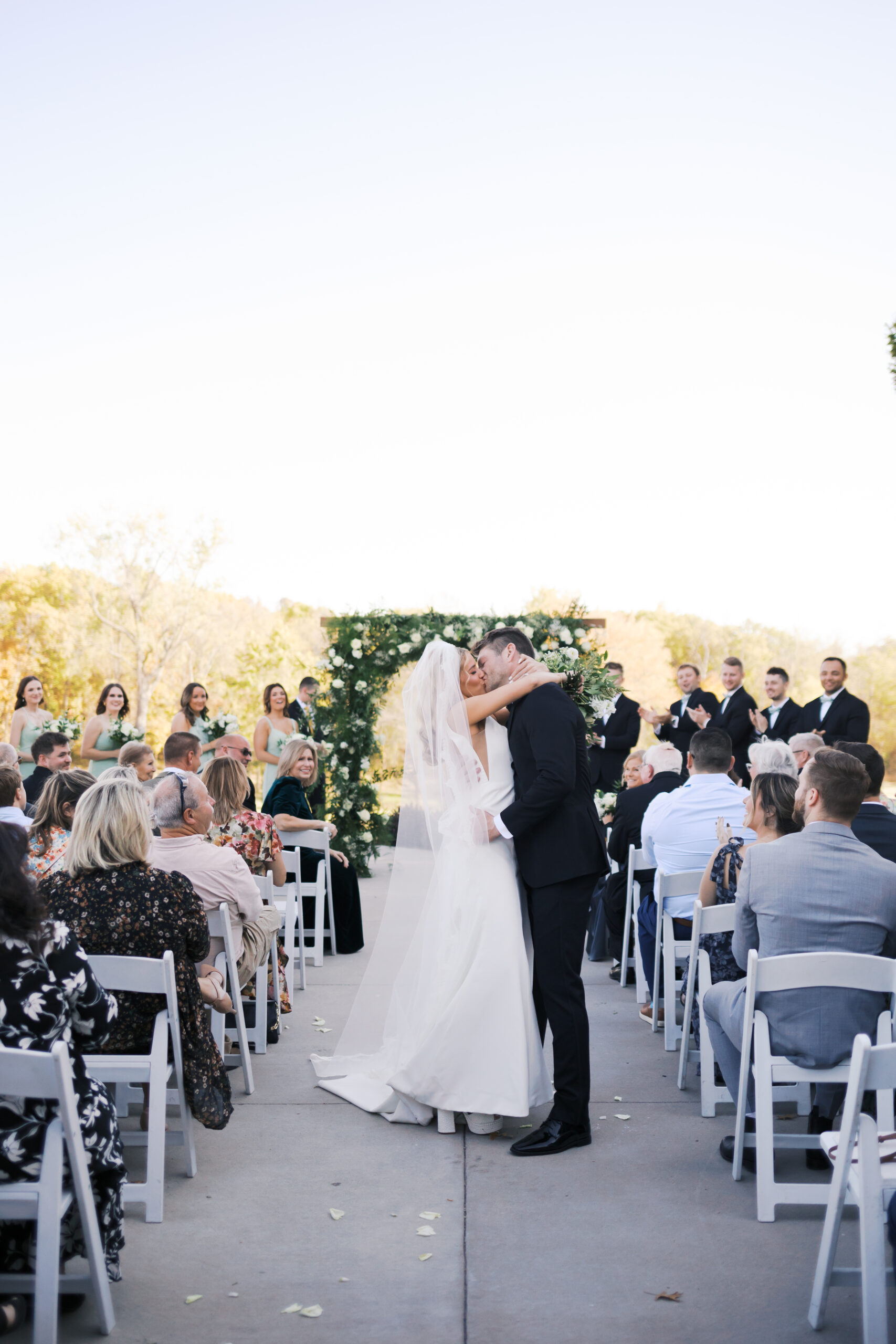Bride and groom share a kiss halfway down the aisle as guests cheer and bridesmaids smile in the background.