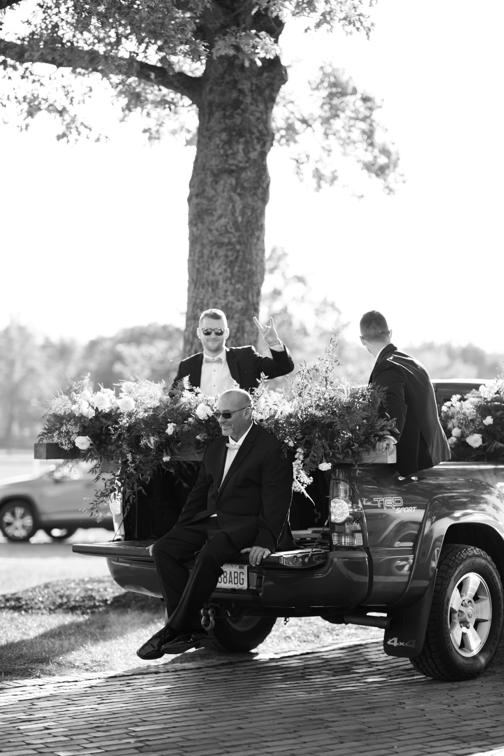 Black and white photo of groomsmen sitting on the bed of a decorated truck filled with floral arrangements, post-ceremony.