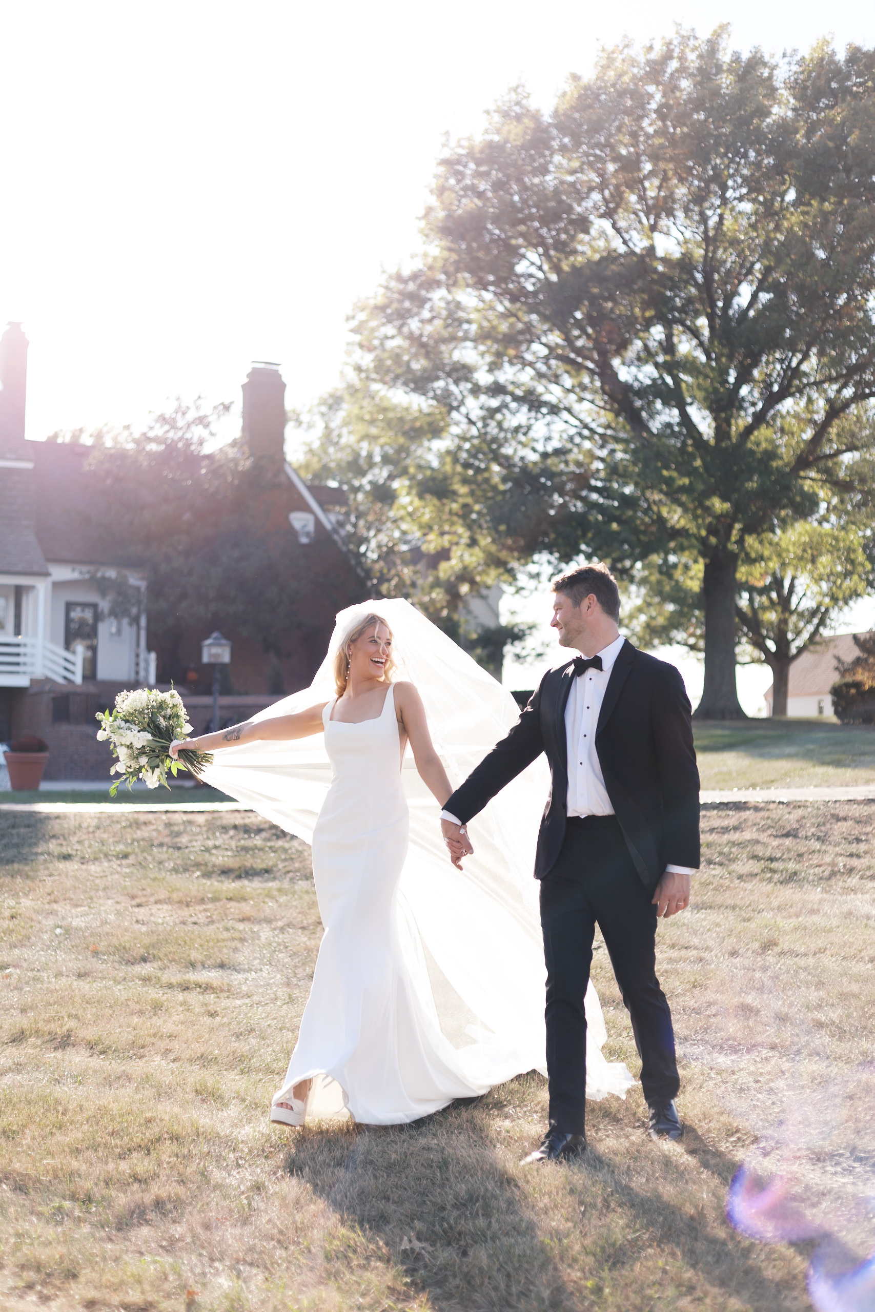 Bride and groom walk hand in hand across a sunlit field, her veil blowing behind her, with Mildale Farm buildings in the background.