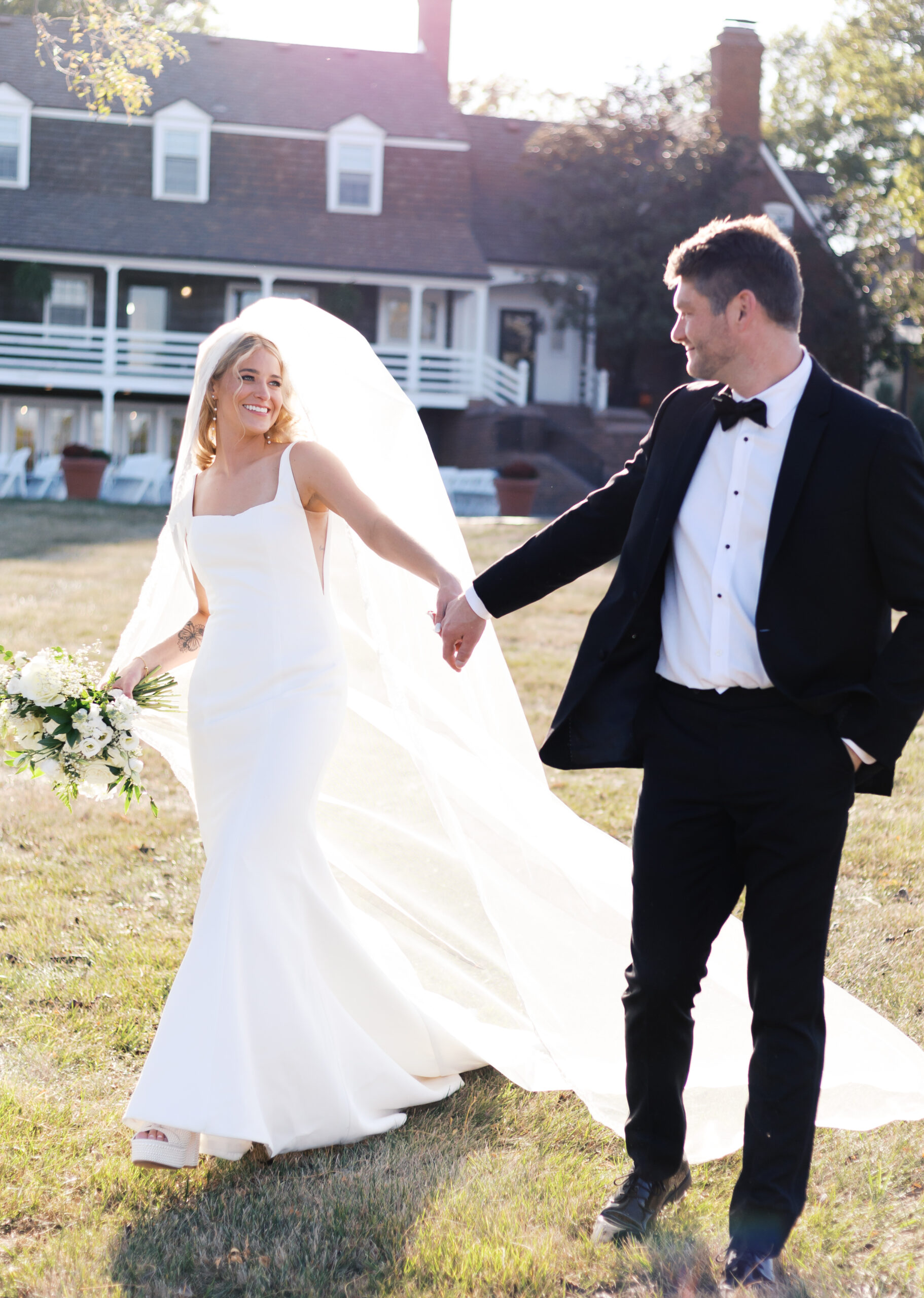 Bride in a white gown and long veil walks hand in hand with the groom in a black tuxedo, smiling in front of Mildale Farm.