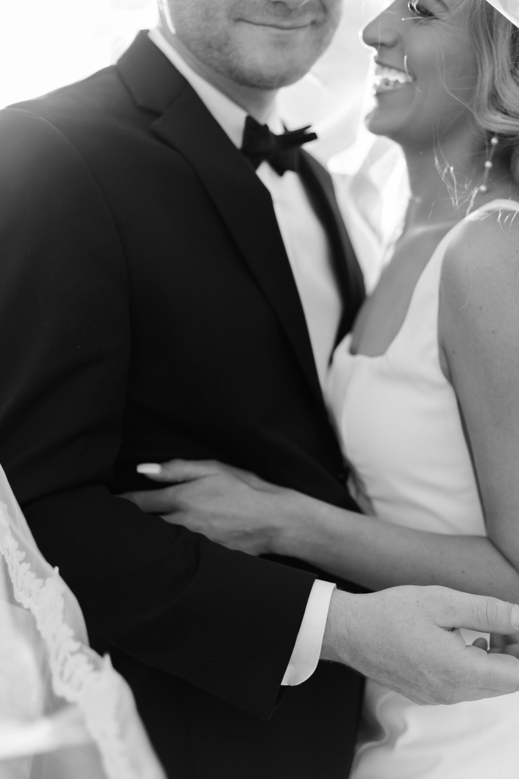 Black and white close-up of bride and groom laughing together, wrapped in her veil during golden hour portraits.