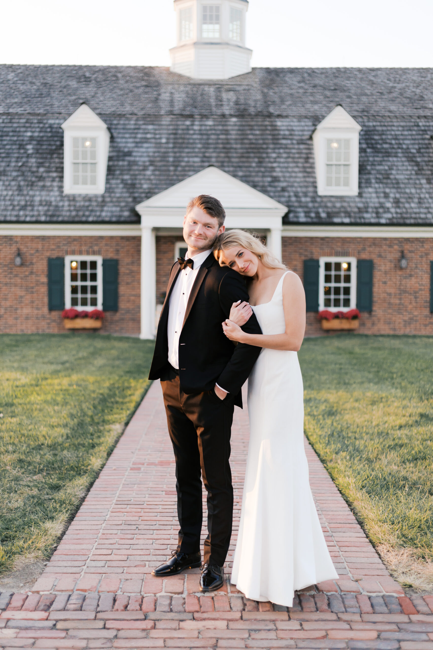 Bride leans her head on the groom’s shoulder as they stand on a brick path in front of Mildale Farm at sunset.