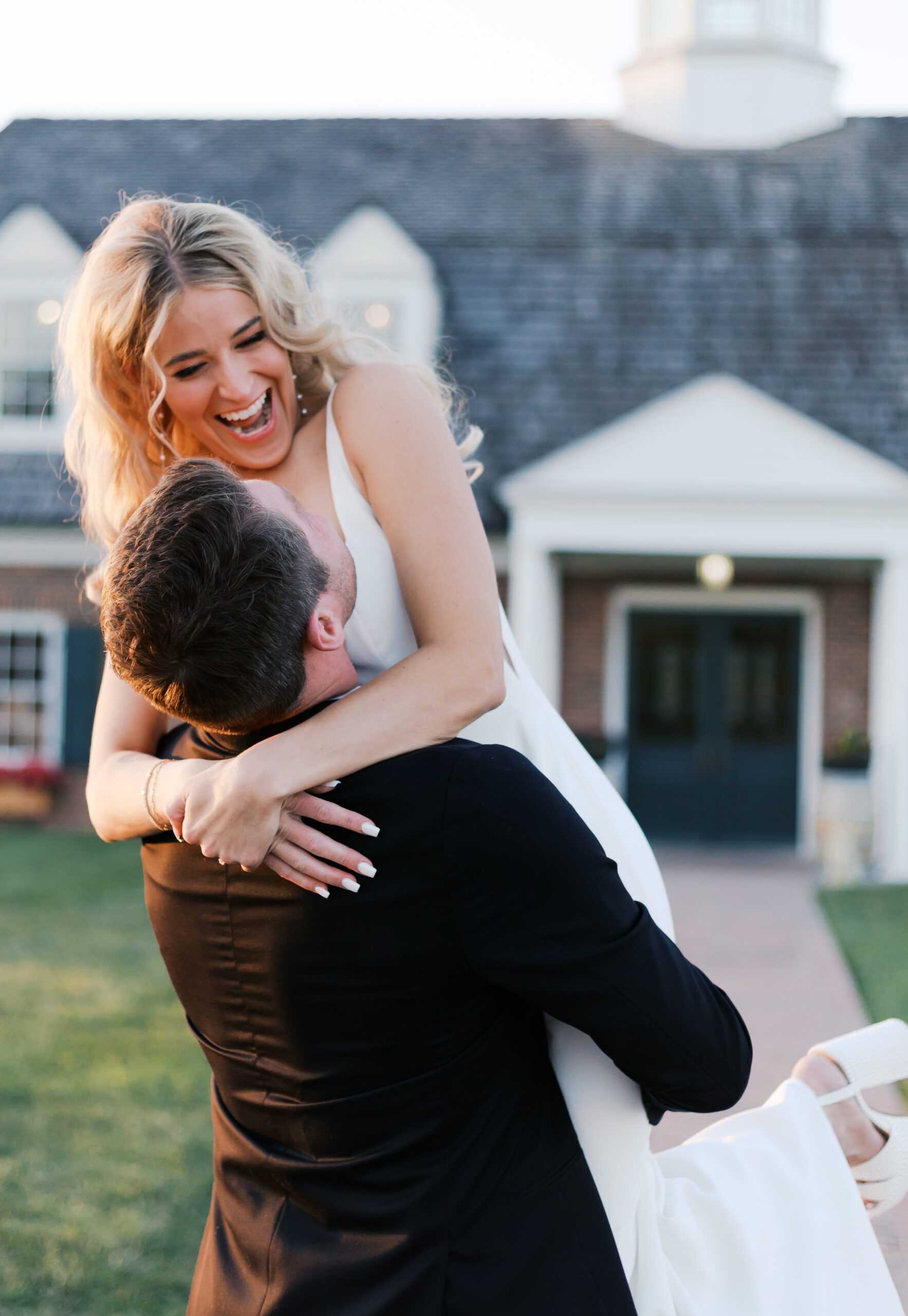 Groom lifts the laughing bride in a white gown, both smiling in front of a brick venue with black shutters and flower boxes.