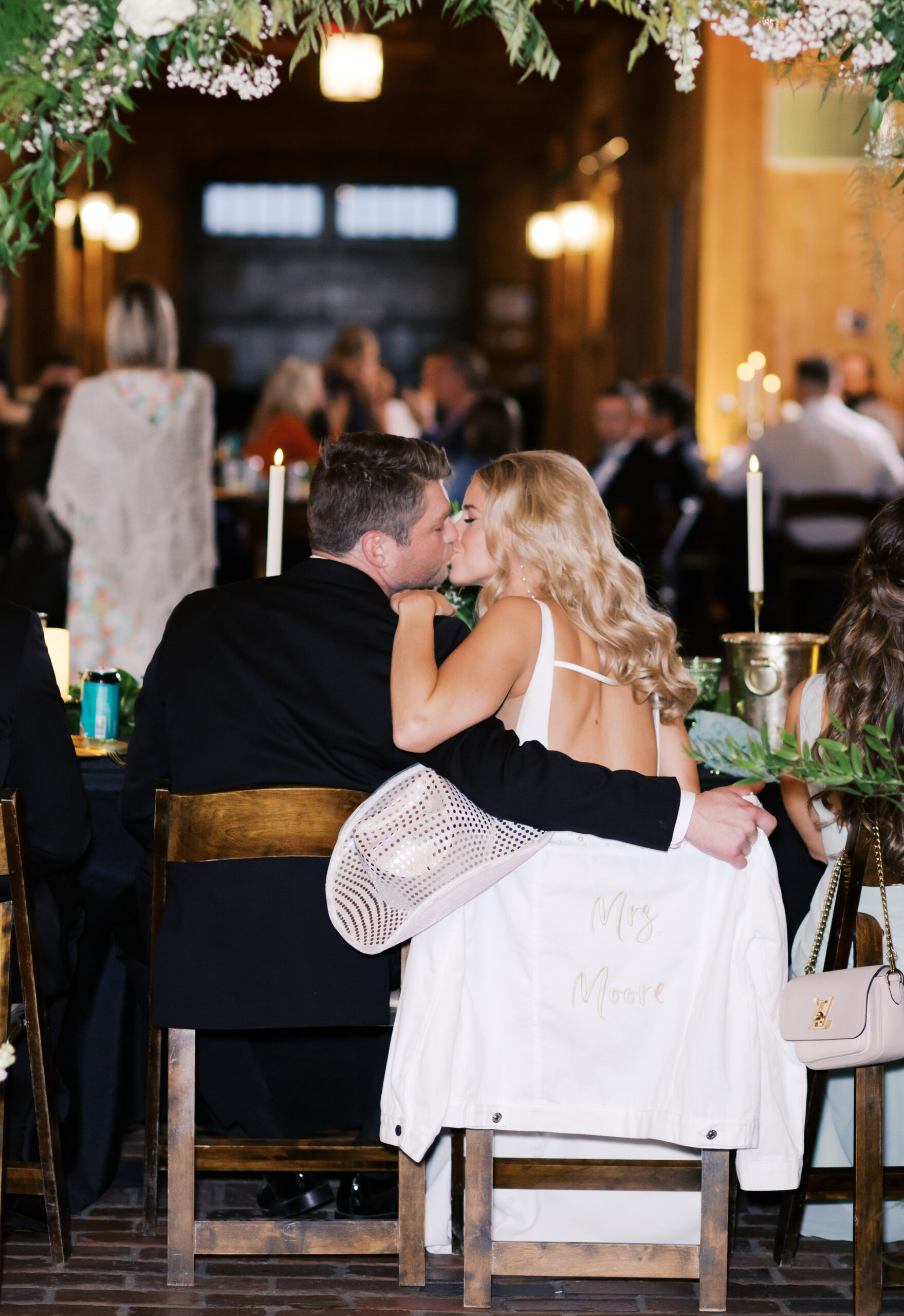 Bride and groom share a kiss at their reception table, her white jacket with “Mrs. Moore” draped over the chair and a rhinestone cowboy hat in hand.