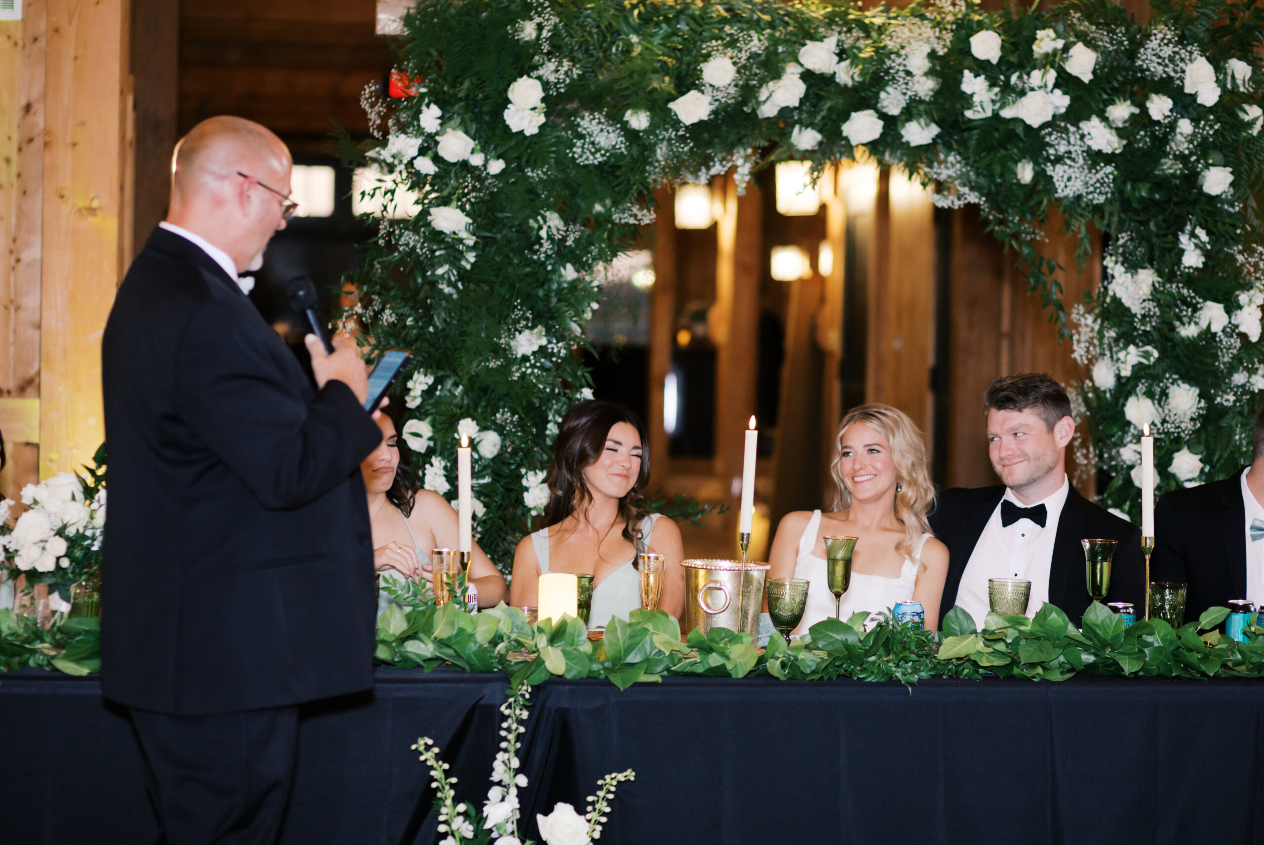 Father of the bride gives a toast during the reception as the bride and groom smile, seated beneath a floral arch of greenery and white rose