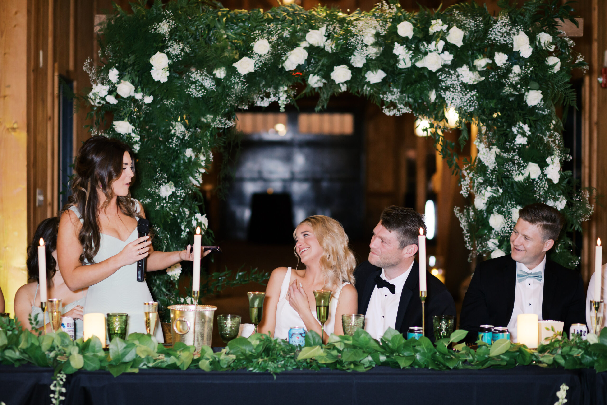 Bridesmaid gives a toast during the reception as the bride and groom smile, seated beneath a floral arch of greenery and white rose