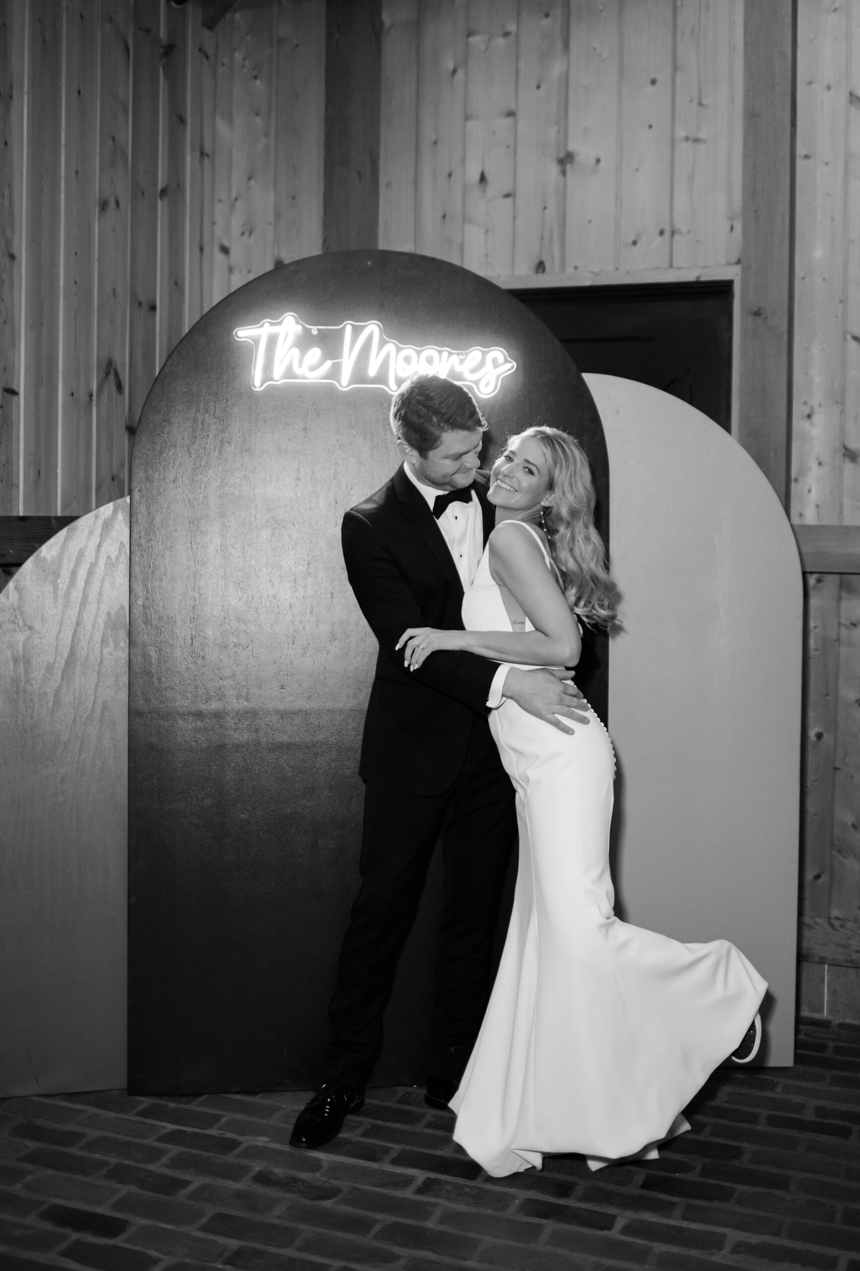 Black and white photo of the married couple embracing in front of a neon sign with The Moore's in script font.