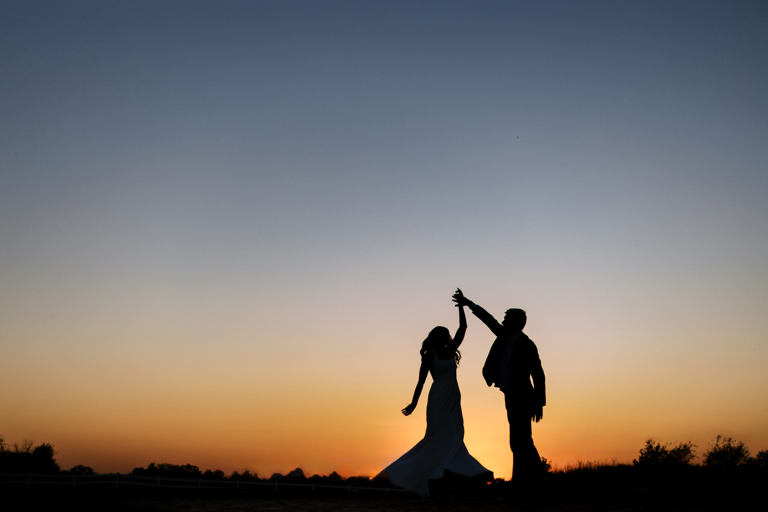 Silhouette portrait of groom twirling bride in front of a setting sun